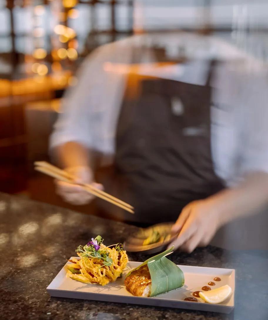A chef plating a tempura and glazed-fish course at Saoke, seen through glass