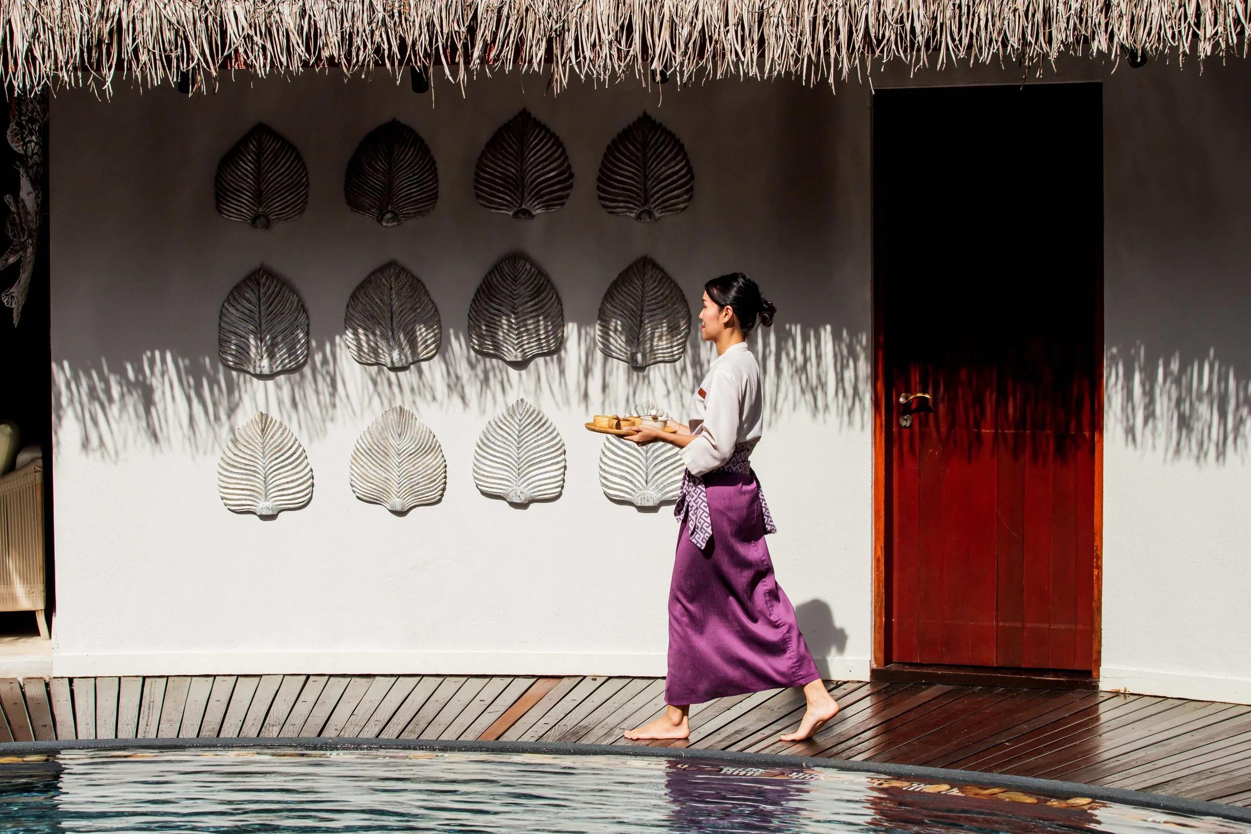 A Devarana Wellness therapist carrying a welcome tray past a reflecting pool at Dusit Thani Maldives