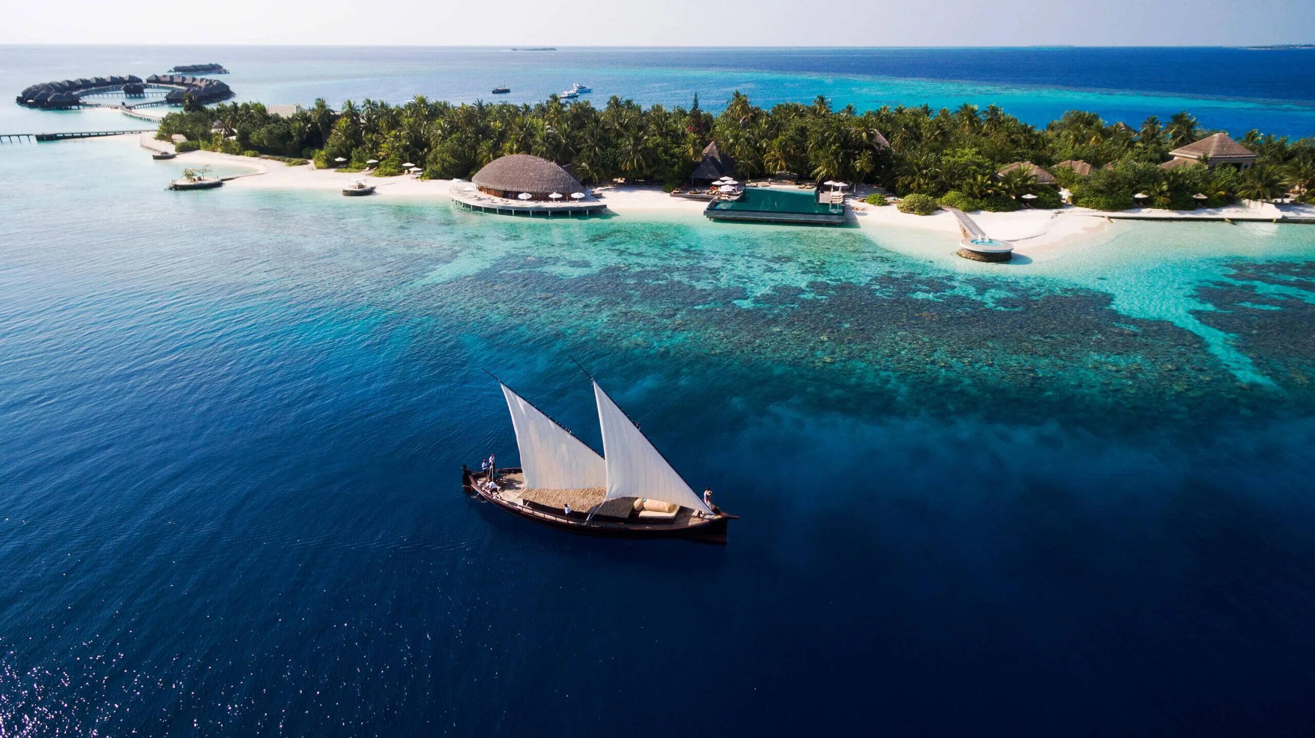 Aerial view of Huvafen Fushi's overwater villas curving around the reef, a traditional dhoni under full sail in the foreground
