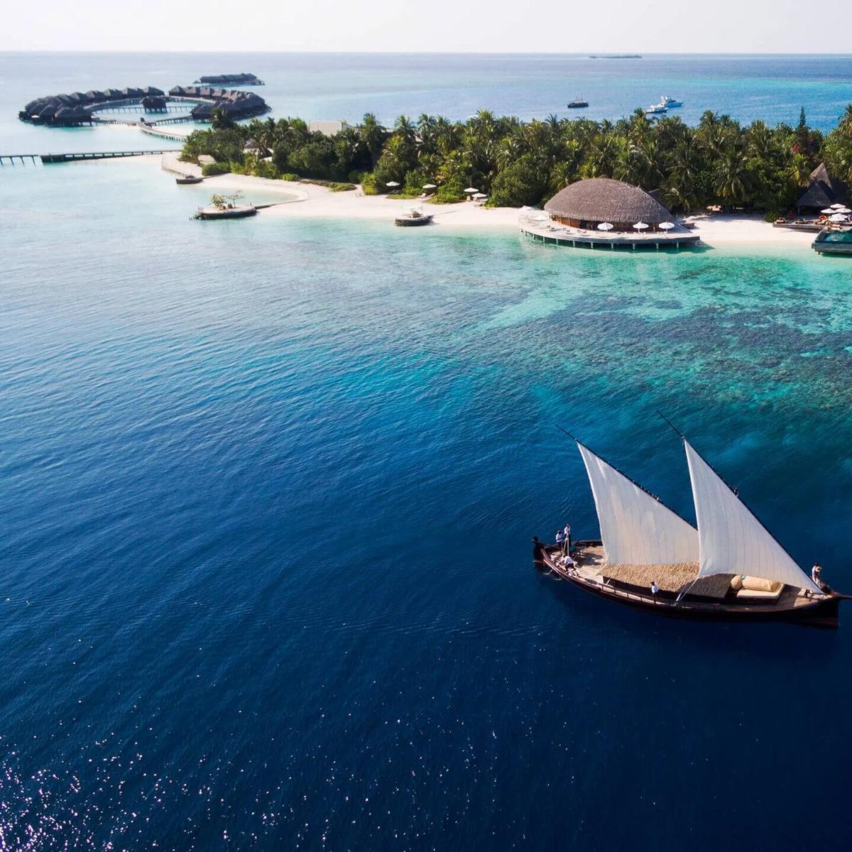 Aerial view of Huvafen Fushi's overwater villas curving around the reef, a traditional dhoni under full sail in the foreground