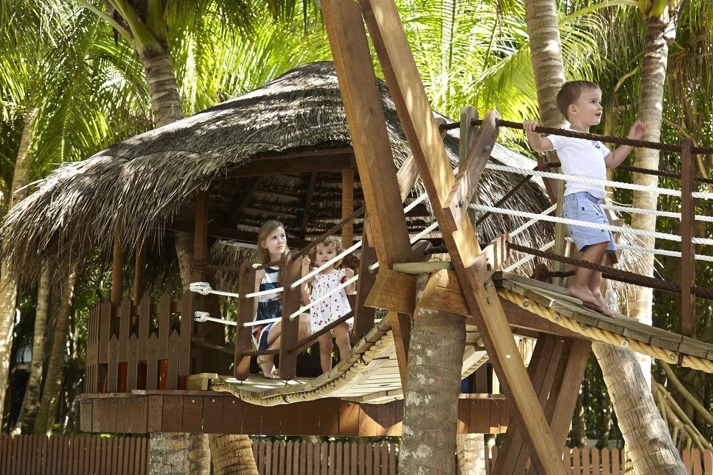 Three children playing on a rope-bridge treehouse structure among palm trees at Dusit Thani Maldives' Kids' Club