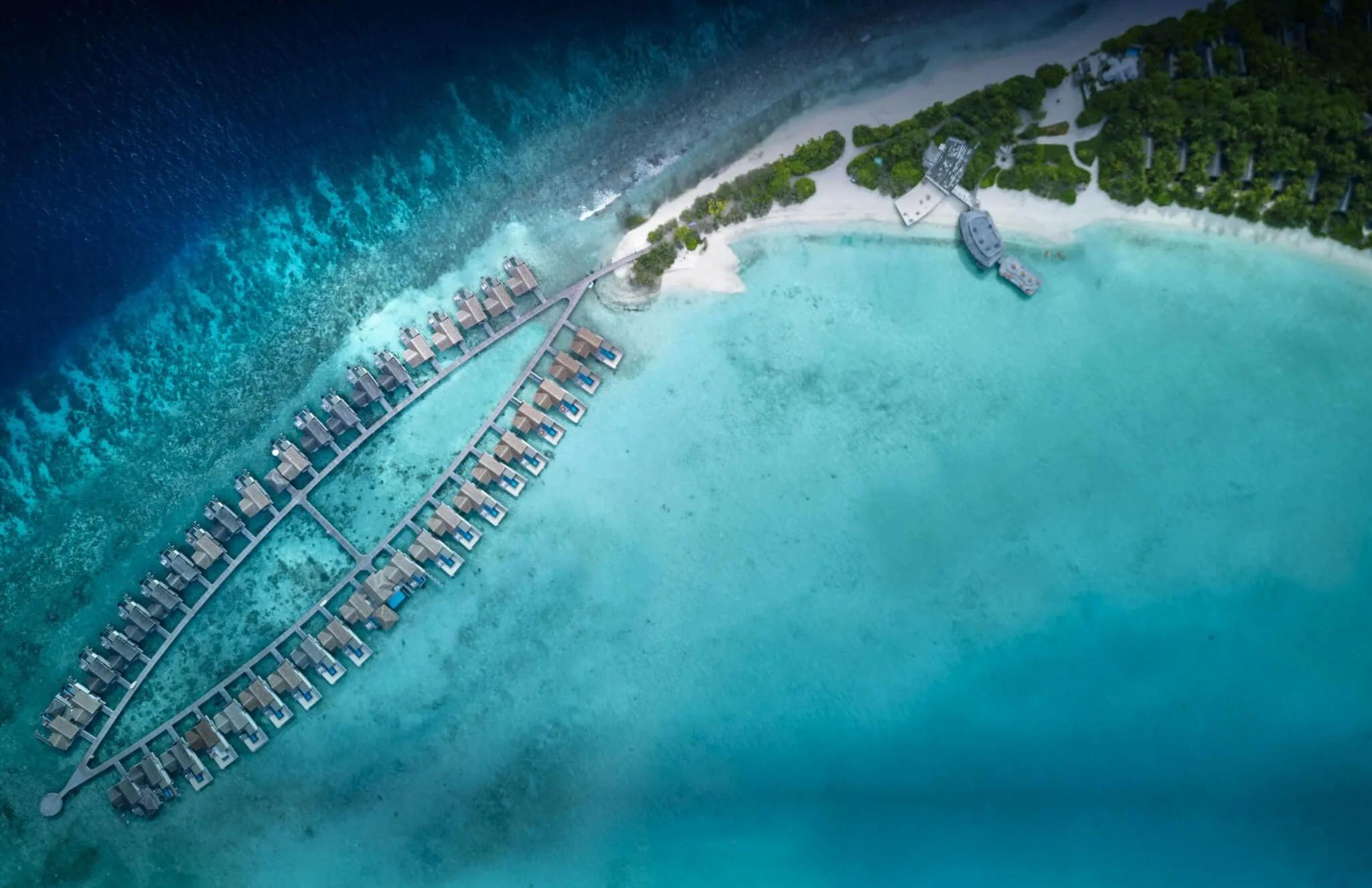 Aerial view of two rows of overwater villas extending from Sirru Fen Fushi's beach into the reef, the island and a jetty visible at the top of frame