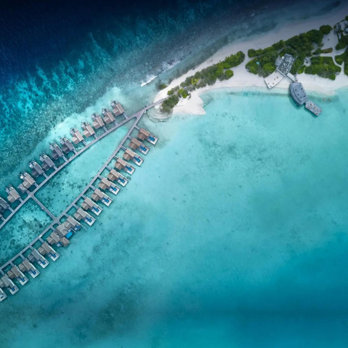 Aerial view of two rows of overwater villas extending from Sirru Fen Fushi's beach into the reef, the island and a jetty visible at the top of frame