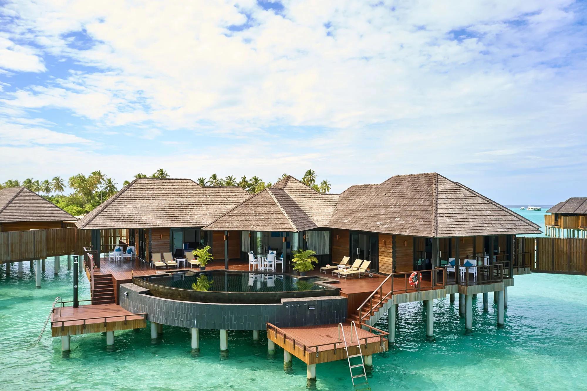 A row of overwater villas seen from the air, turquoise lagoon on both sides and the palm-lined island behind