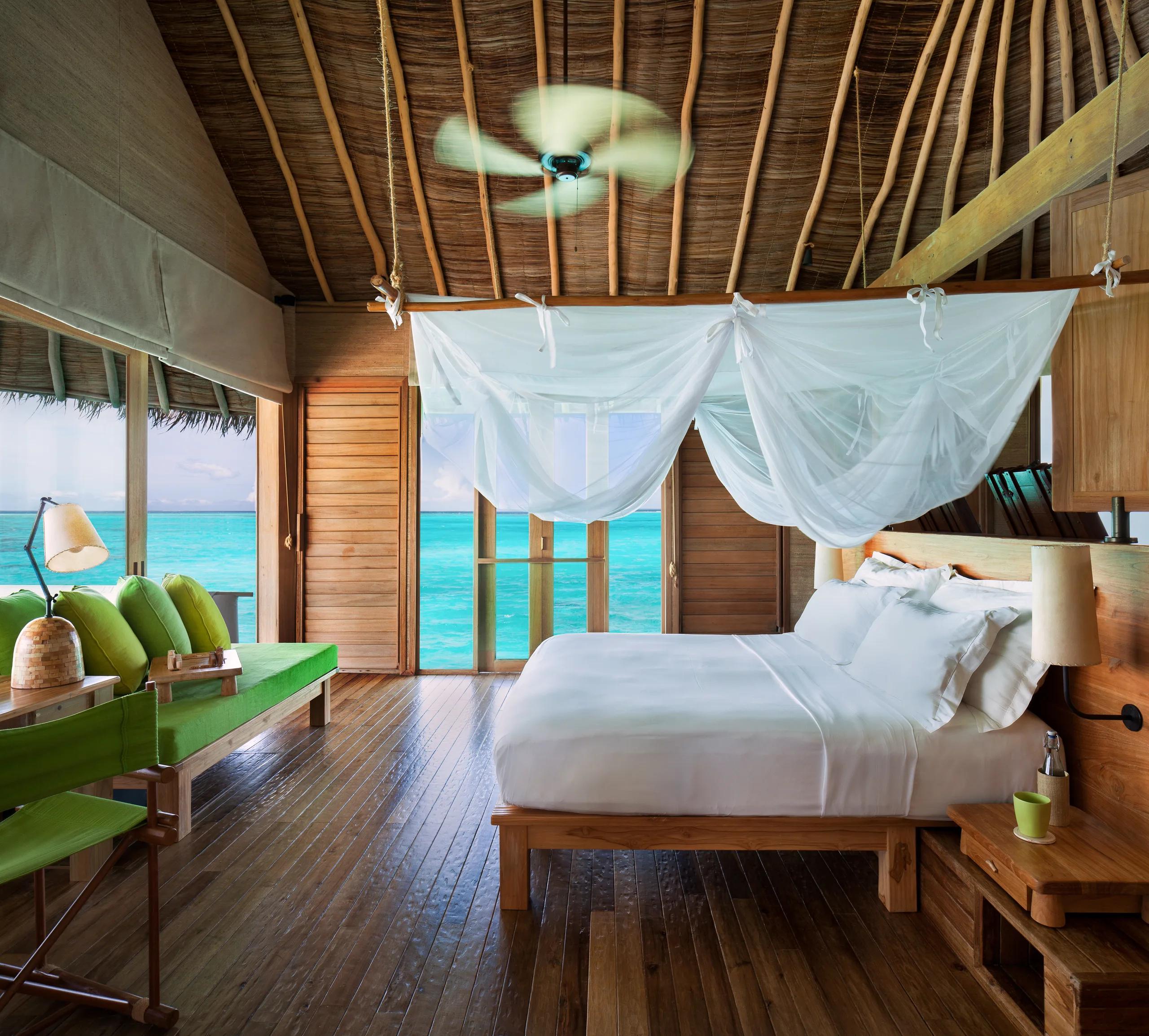 A bedroom in an overwater villa with a sheer canopy over the bed and glass doors opening onto the lagoon