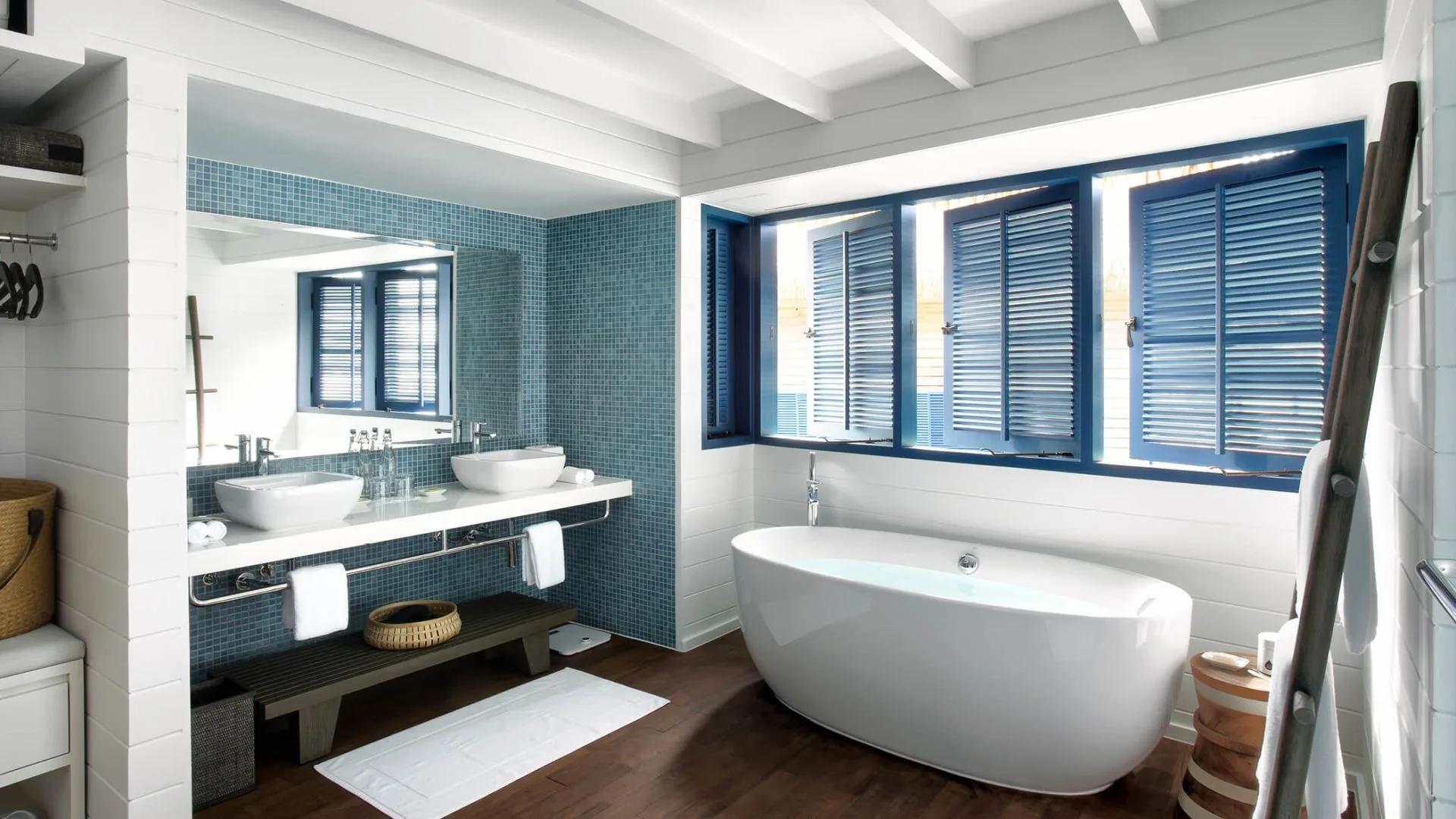 The bathroom of a Dhoni Loft Water Villa, with a freestanding tub set beneath tall blue-shuttered windows