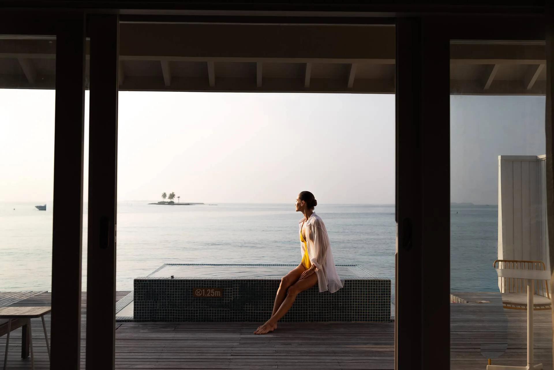 A guest in a yellow swimsuit sitting at the edge of an Ocean Overwater Villa's plunge pool at dusk, a small island visible on the horizon