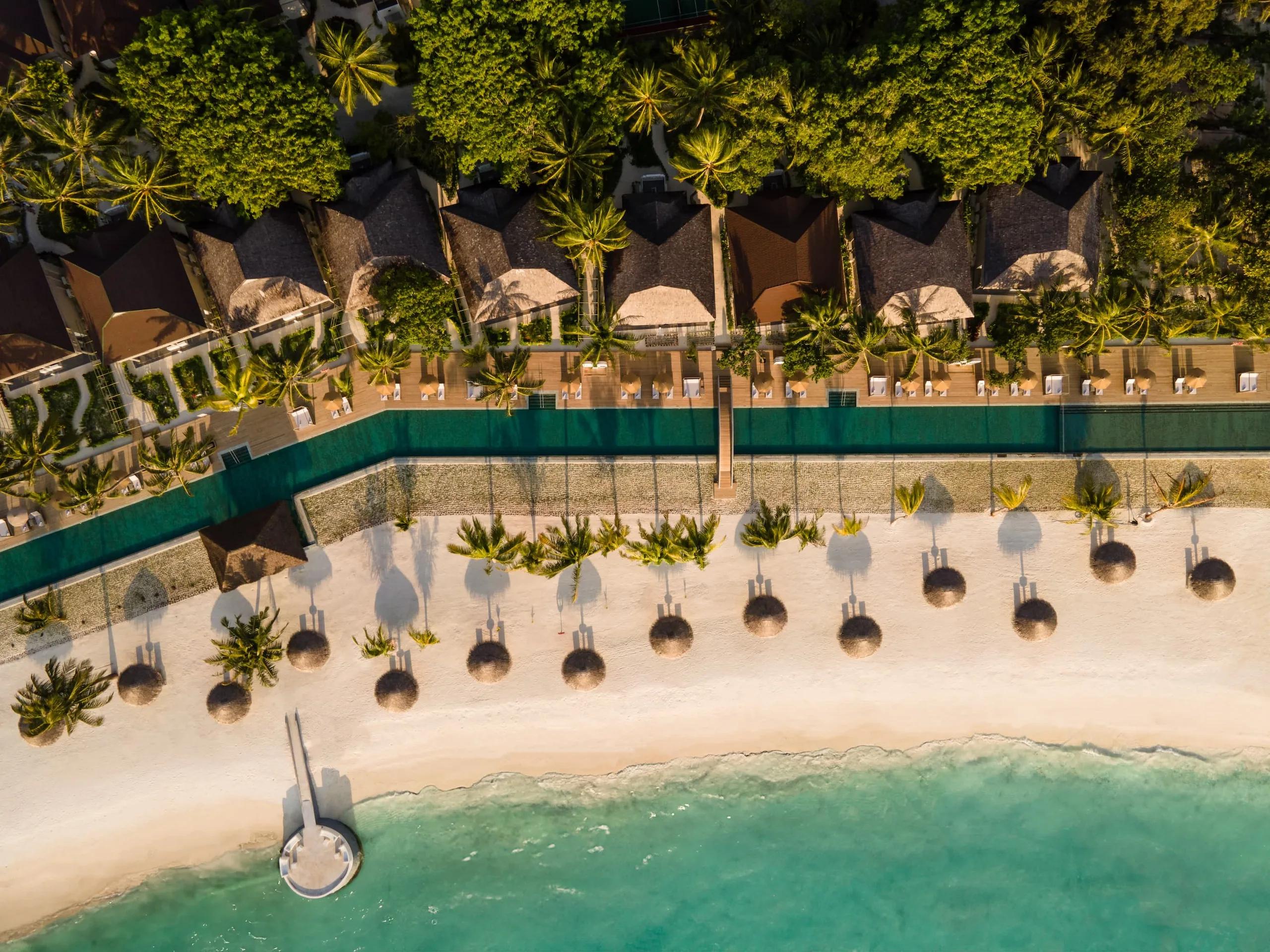 Aerial view at golden hour of an overwater pool deck lined with sun loungers and umbrellas, set between beach villas and the lagoon at Sun Siyam Olhuveli