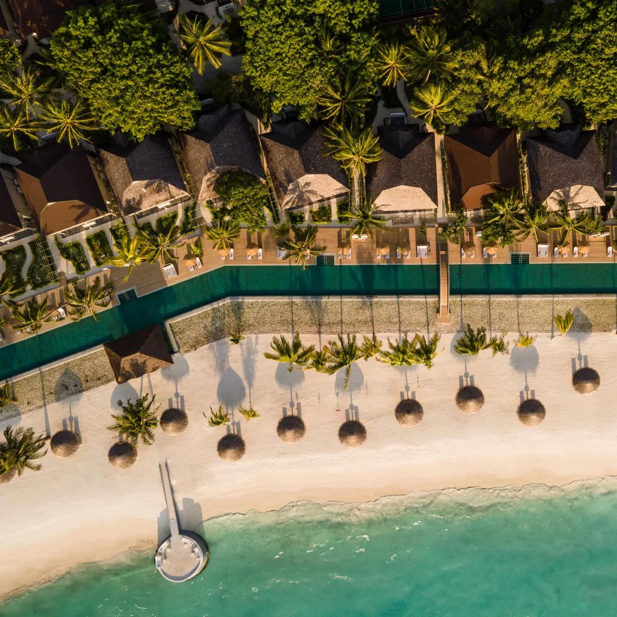 Aerial view at golden hour of an overwater pool deck lined with sun loungers and umbrellas, set between beach villas and the lagoon at Sun Siyam Olhuveli