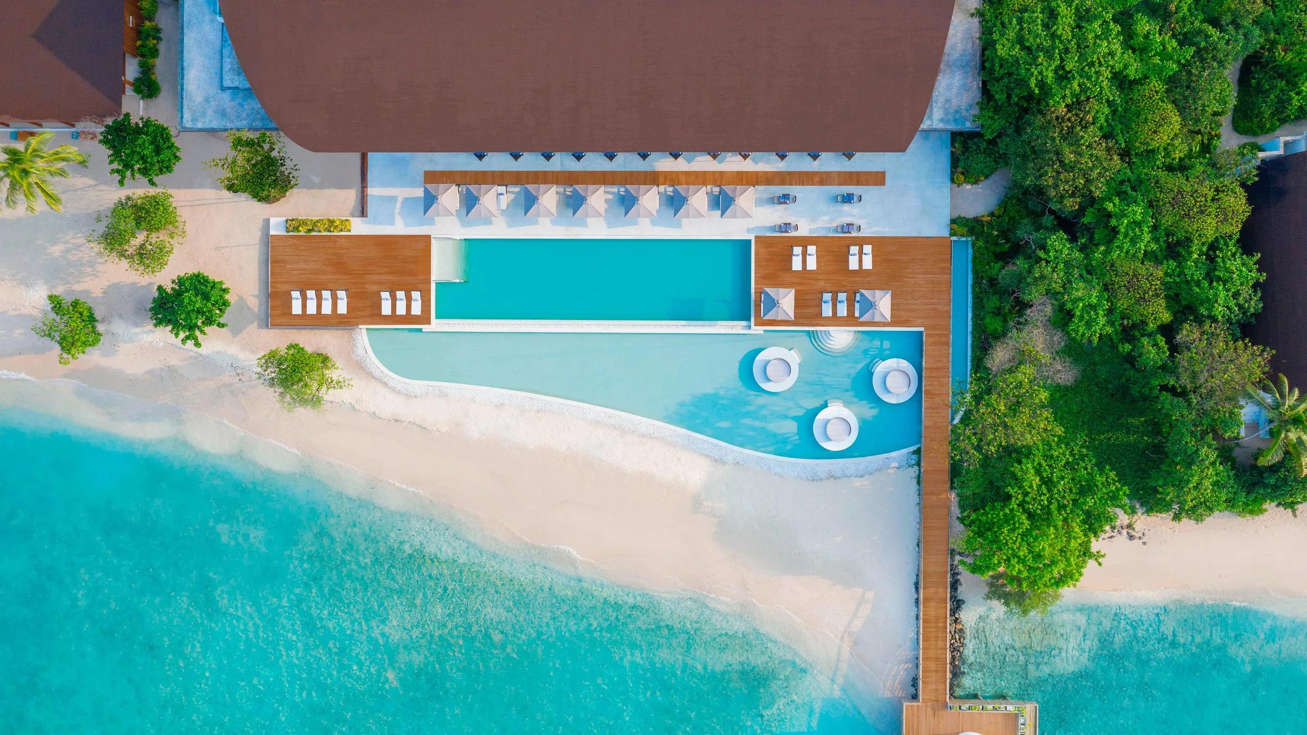 A top-down view of the resort's main pool and beach, decked terraces on one side and open sand on the other