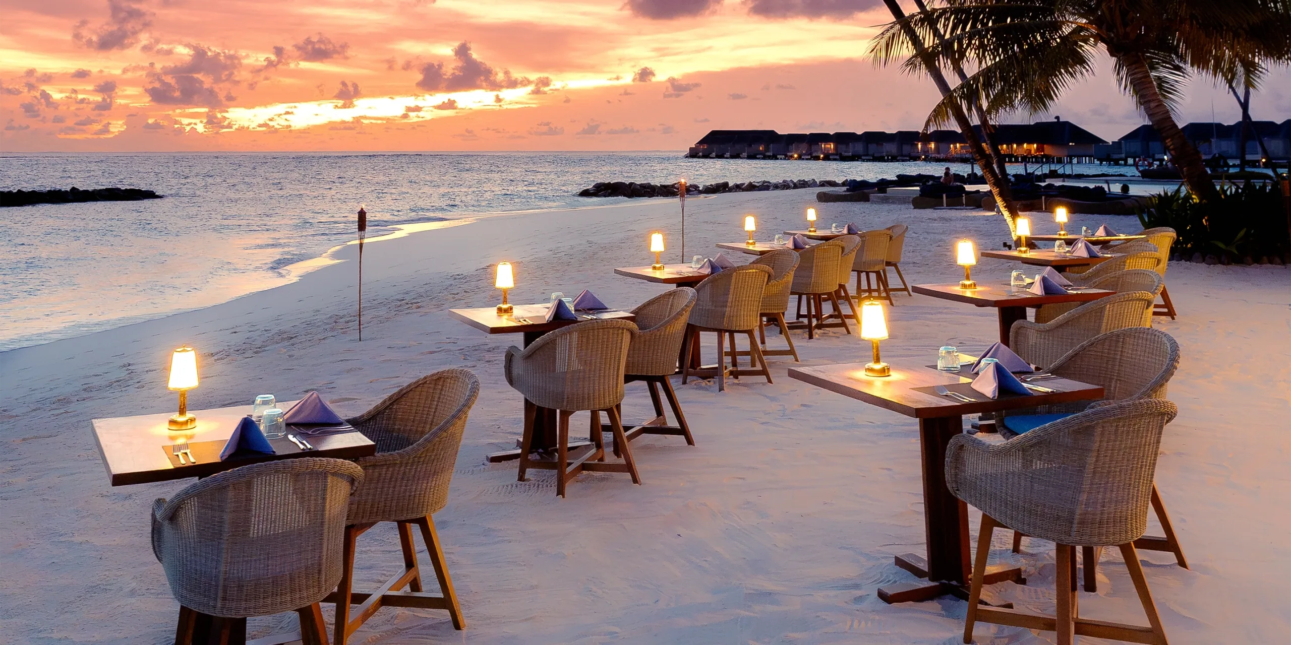 Fuego's beachfront barbecue tables lit for dinner as the sun sets behind the overwater villas at Baglioni Maldives
