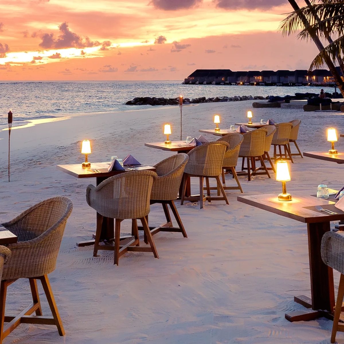 Fuego's beachfront barbecue tables lit for dinner as the sun sets behind the overwater villas at Baglioni Maldives