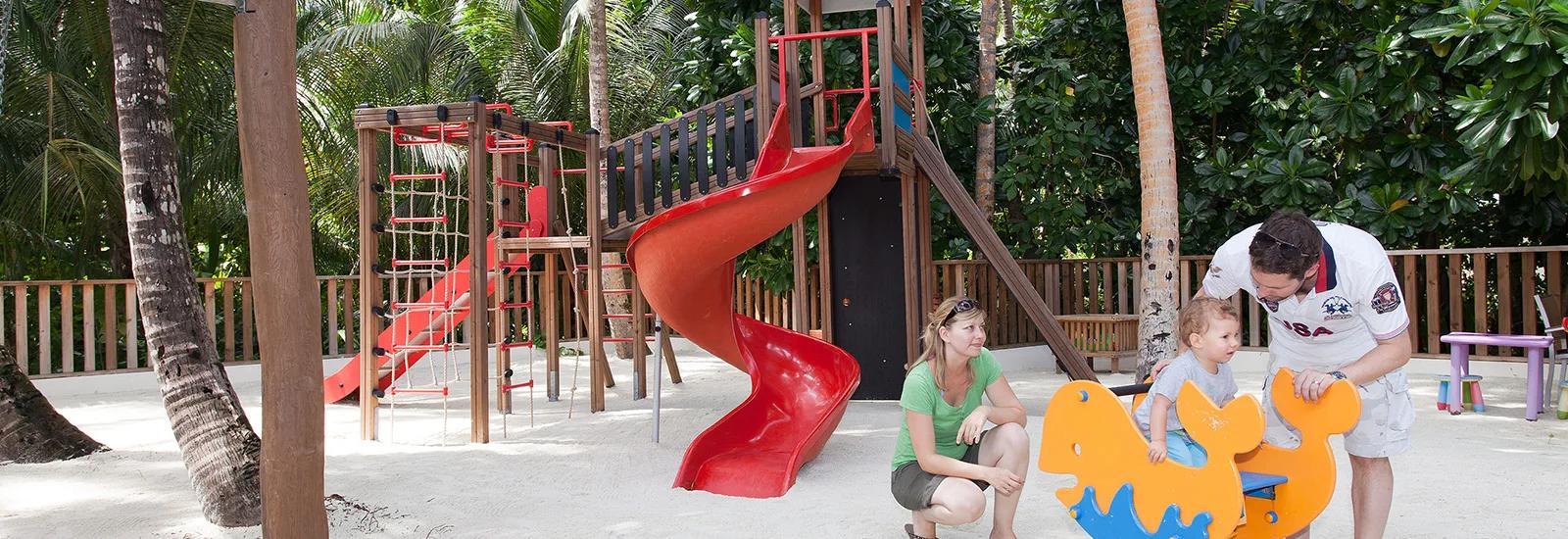 A toddler on a spring rocker at Heron Kids Club's outdoor play area, a red slide and climbing frame behind, supervised by two adults