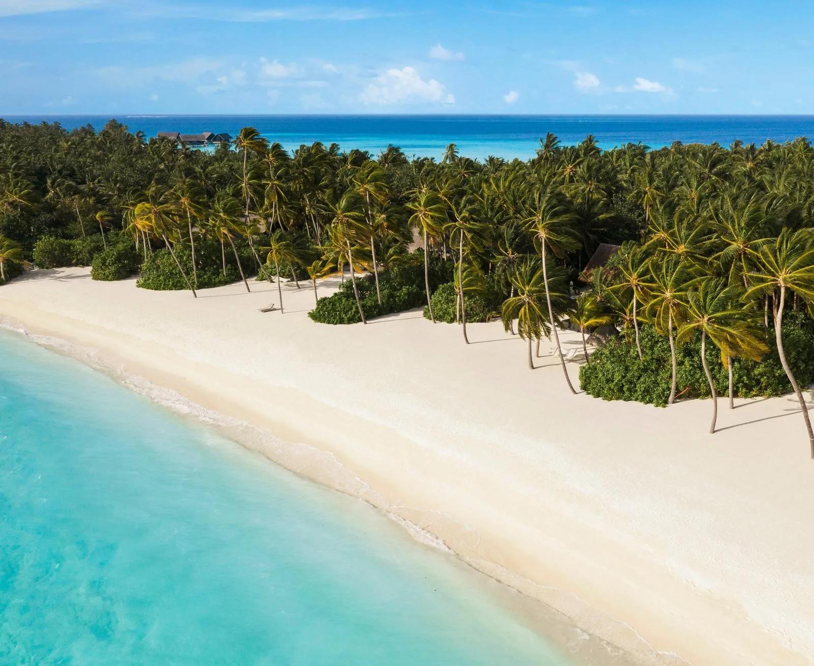 Aerial view of a white-sand beach lined with palm trees at One&Only Reethi Rah, overwater villas visible in the lagoon beyond