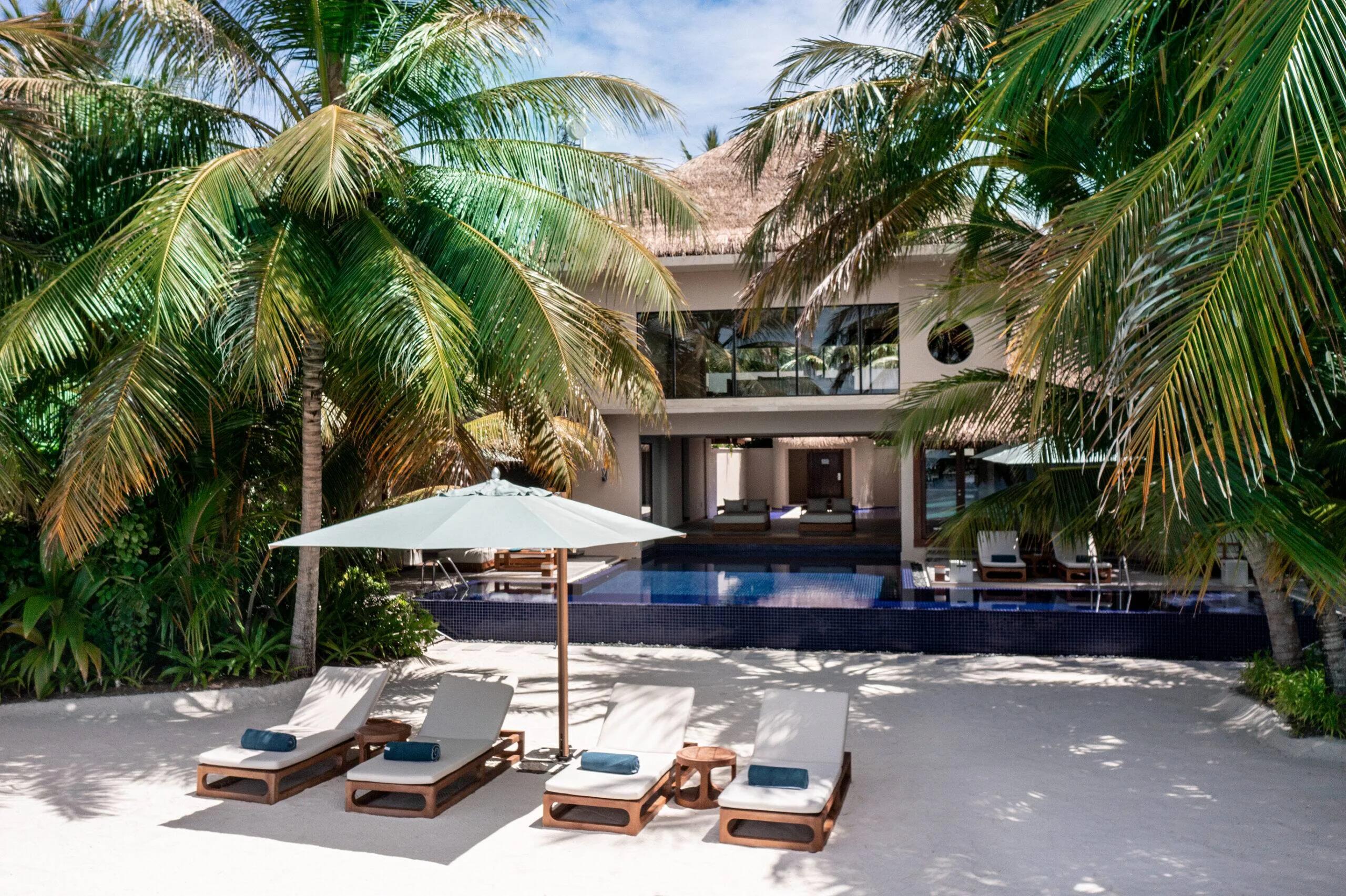 The Three-Bedroom Beach Pavilion behind palm trees, its two-storey structure and infinity pool visible past a row of beach loungers