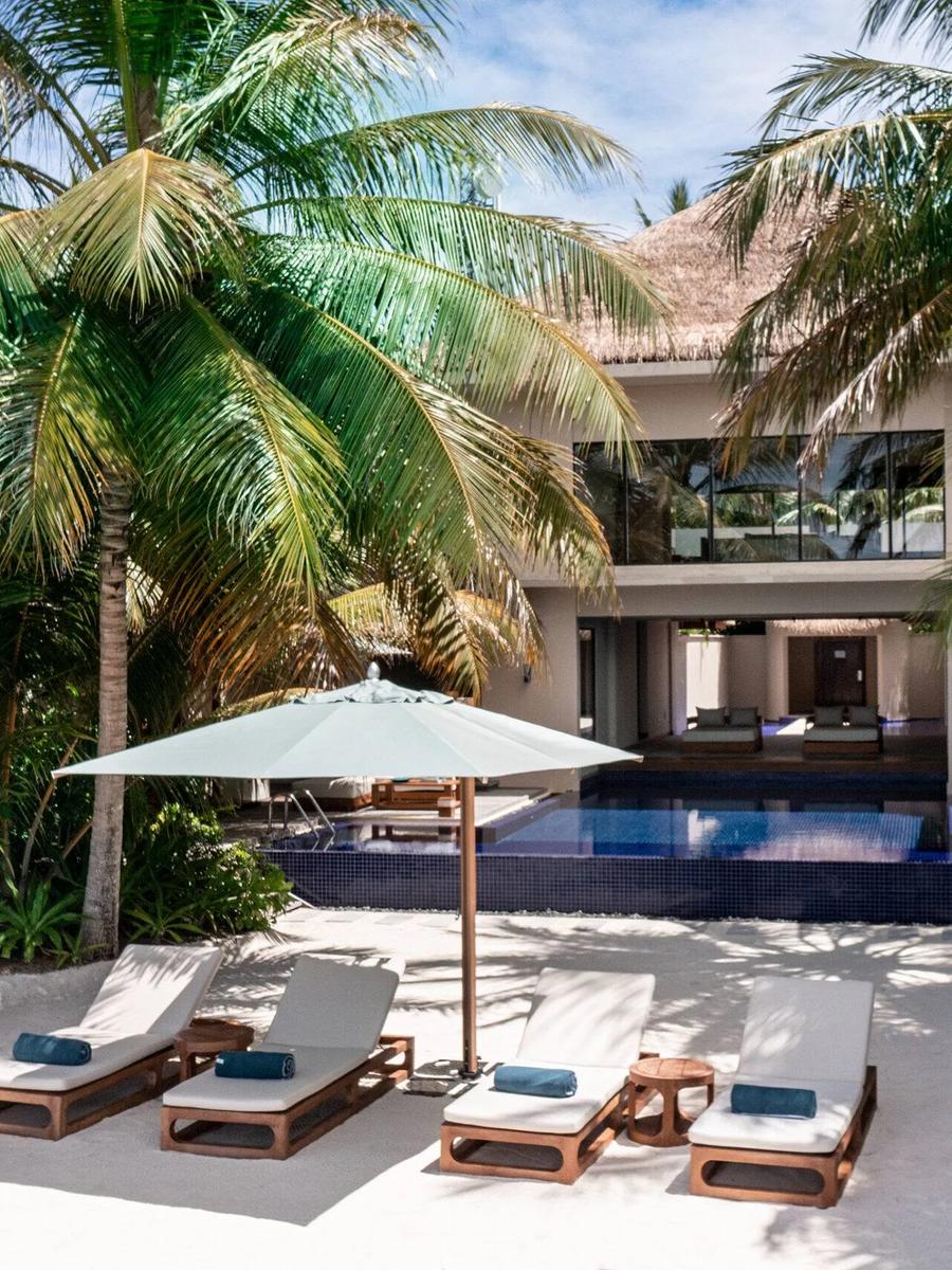 The Three-Bedroom Beach Pavilion behind palm trees, its two-storey structure and infinity pool visible past a row of beach loungers