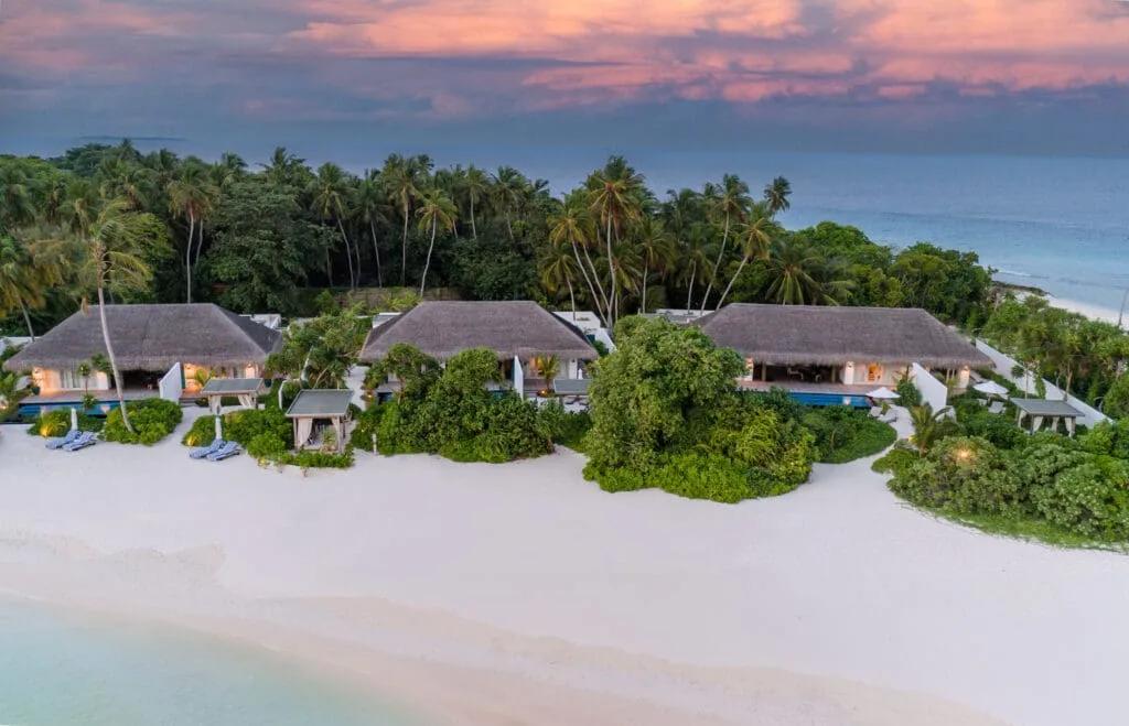 Three thatched-roof beachfront villas set side by side along the sand at dusk, palm trees and ocean behind