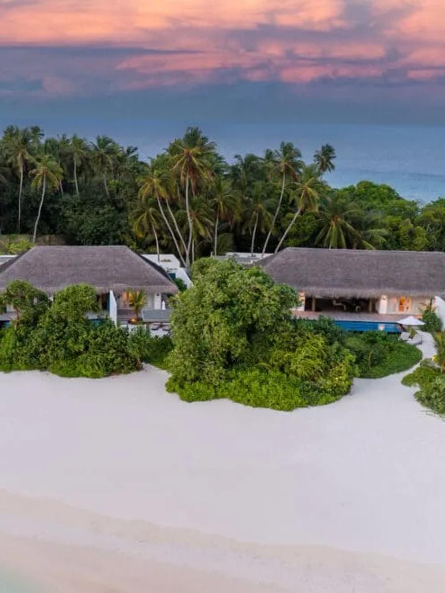 Three thatched-roof beachfront villas set side by side along the sand at dusk, palm trees and ocean behind