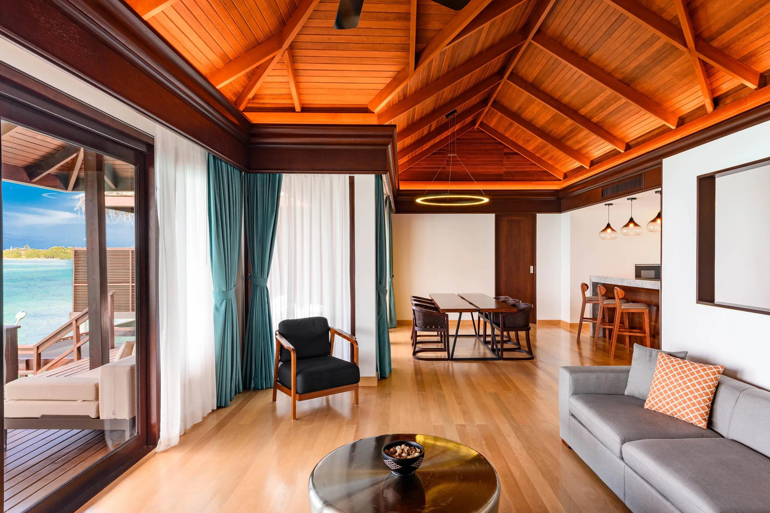 Living and dining area of the Two-Bedroom Overwater Villa With Pool, with a dining table, sofa and breakfast bar under a vaulted wood ceiling