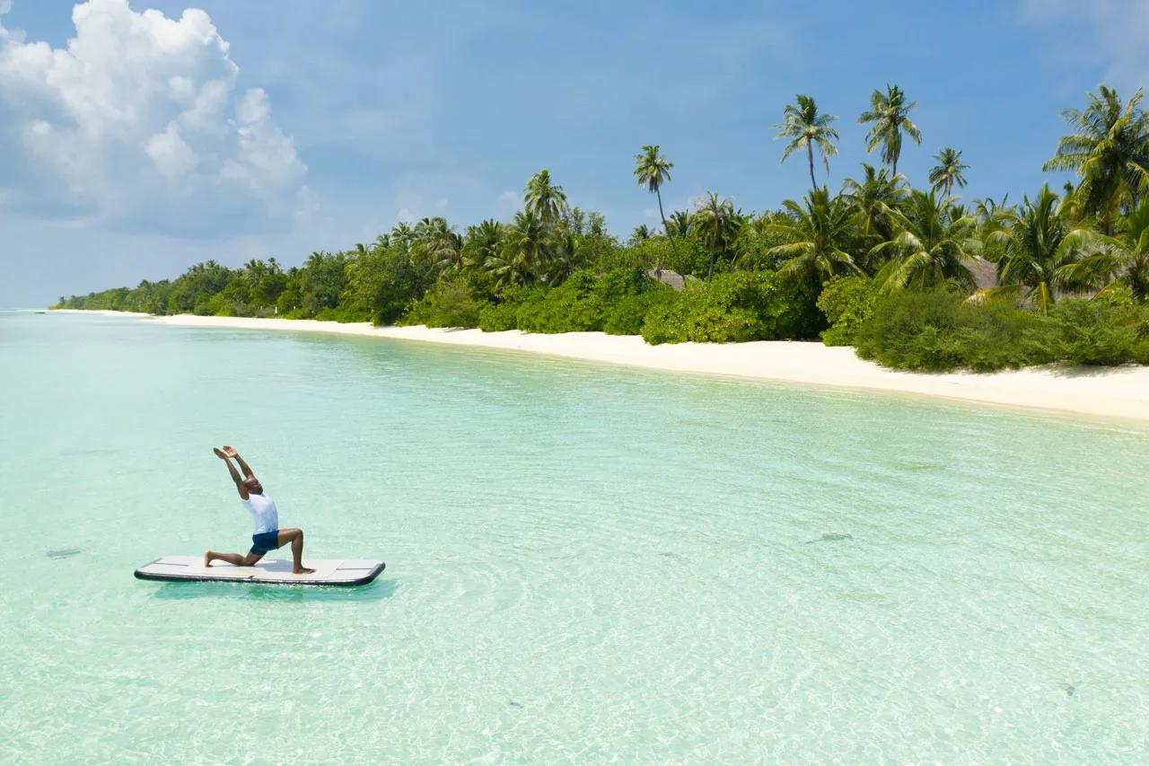 A guest holding a yoga pose on a paddleboard in the shallows off the beach, palm-lined shore behind