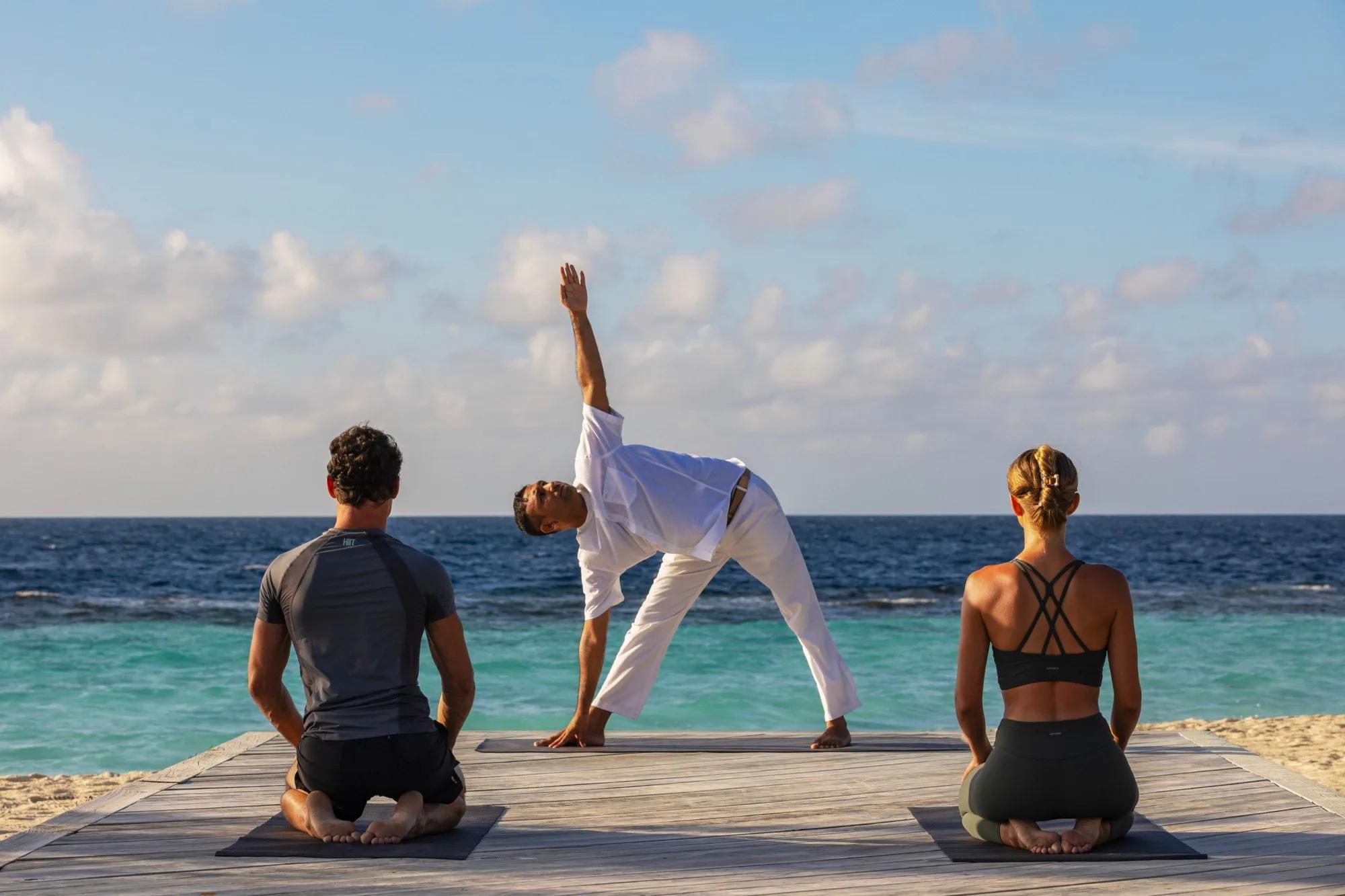 A yoga instructor in white leading a triangle-pose stretch on a jetty at sunrise, two guests seated on mats behind him with the open ocean ahead