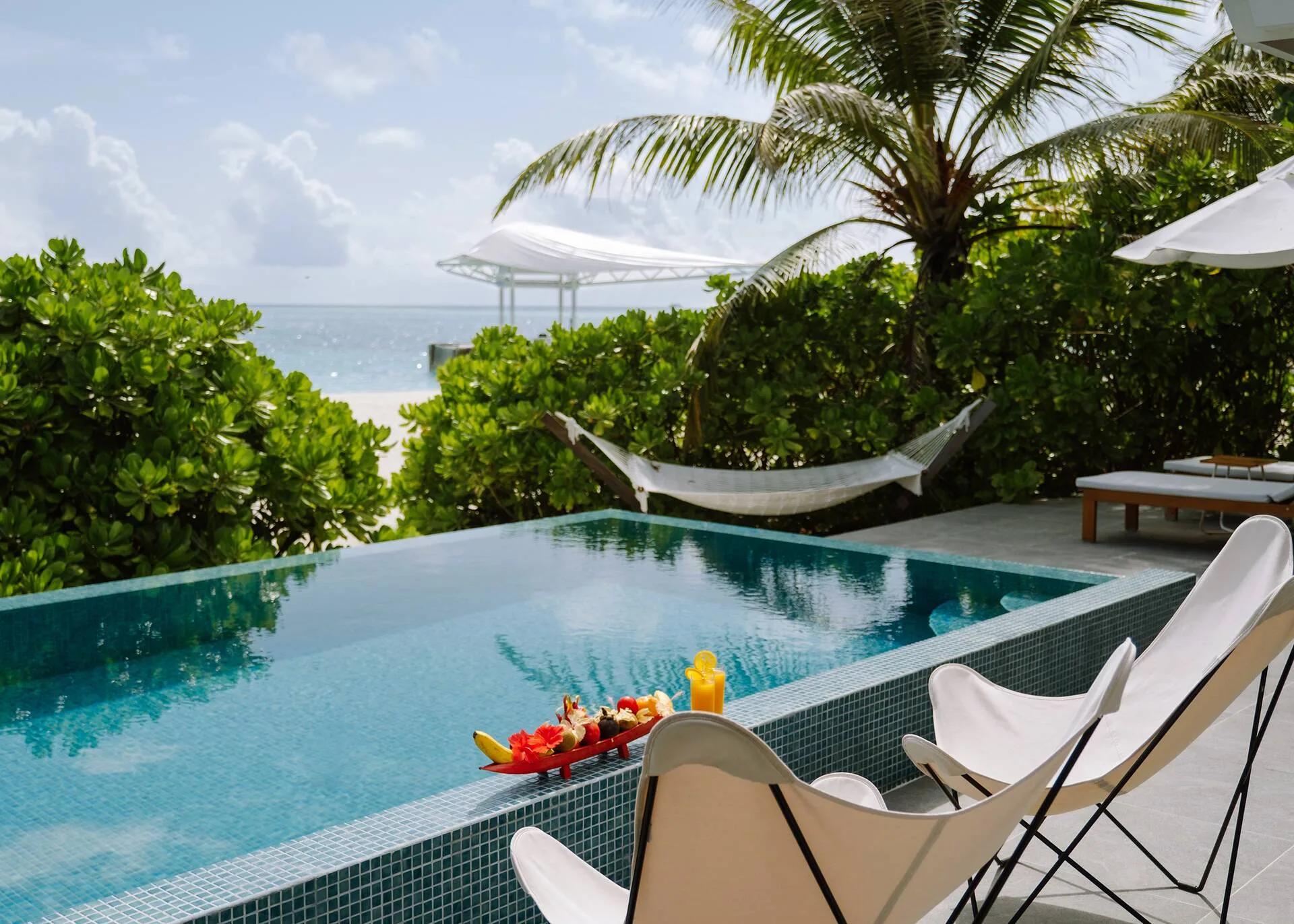 A private pool deck at a Two Bedroom Ocean Beach Villa with a hammock and a fruit-topped breakfast tray poolside, the beach beyond