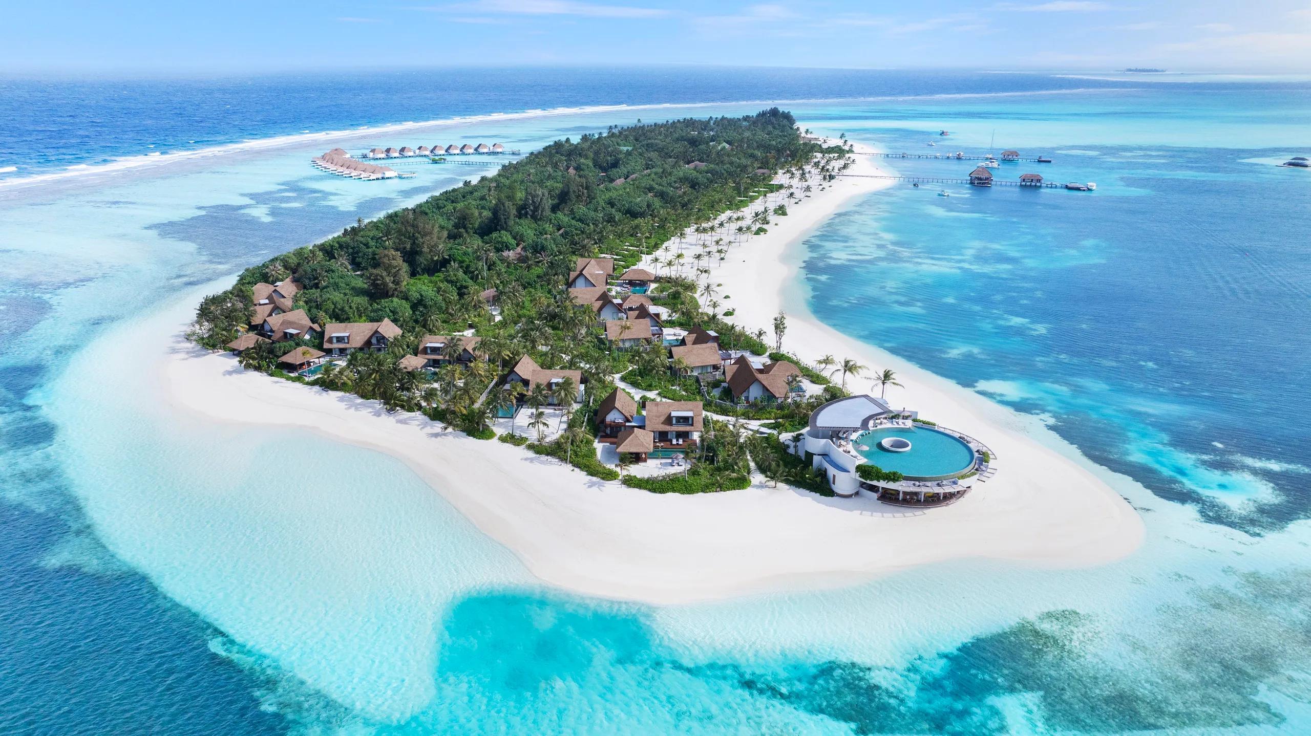 Aerial view of Six Senses Kanuhura's point, with a round infinity pool, thatched villas among palm trees, and overwater villas extending into the lagoon on both sides.