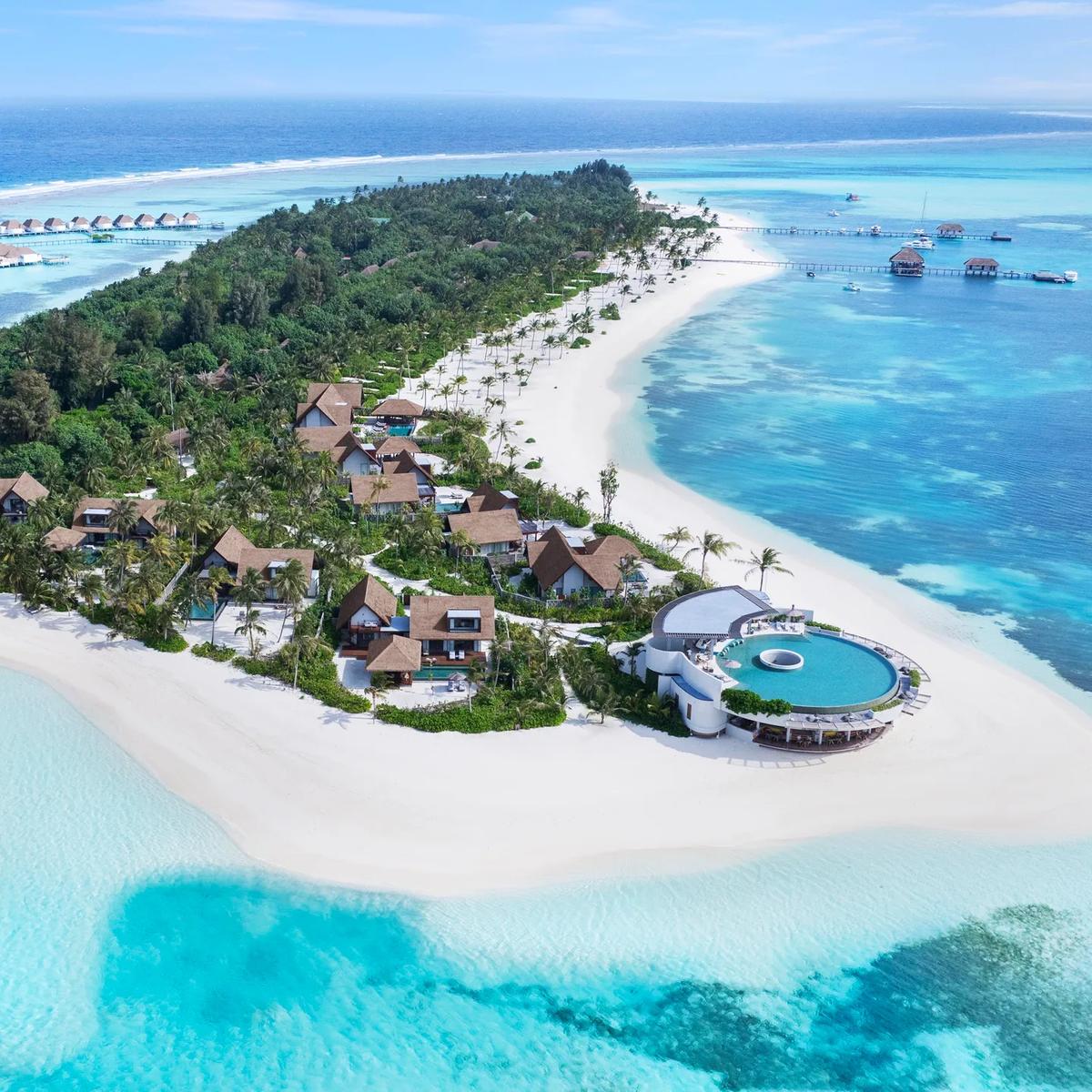 Aerial view of Six Senses Kanuhura's point, with a round infinity pool, thatched villas among palm trees, and overwater villas extending into the lagoon on both sides.