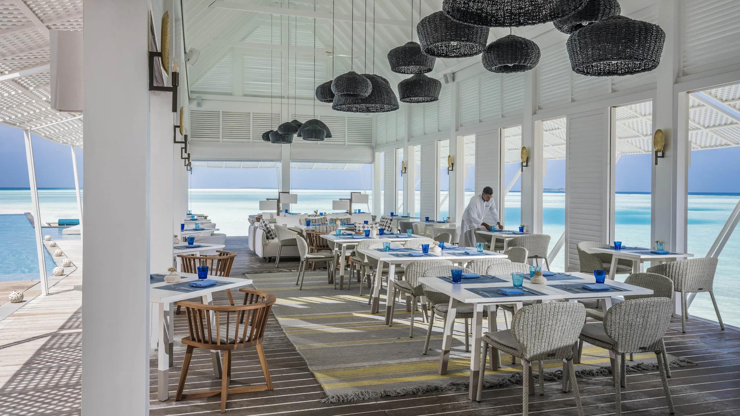 Blu's overwater dining room set for service under woven pendant lamps, with a server arranging a table beside the lagoon