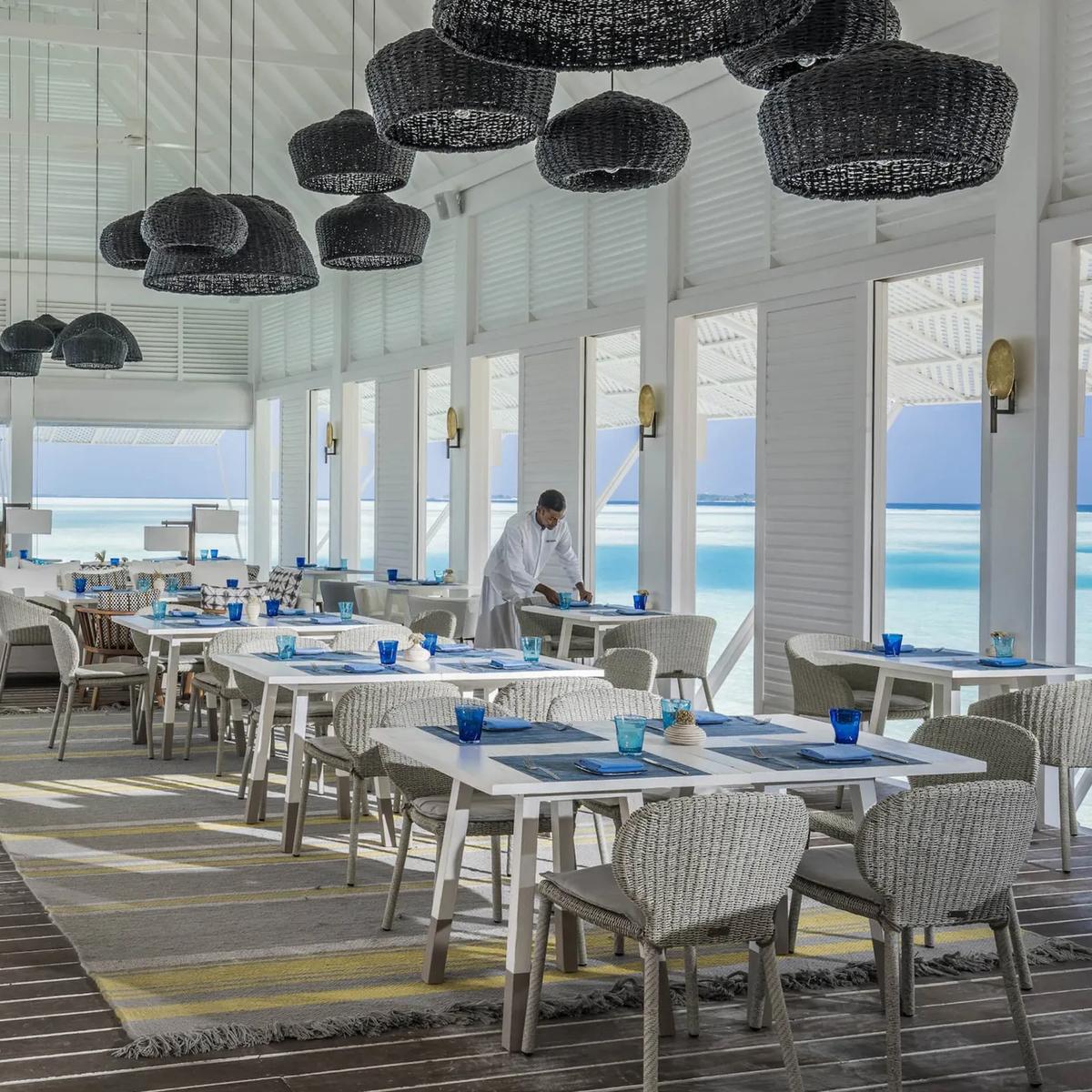 Blu's overwater dining room set for service under woven pendant lamps, with a server arranging a table beside the lagoon