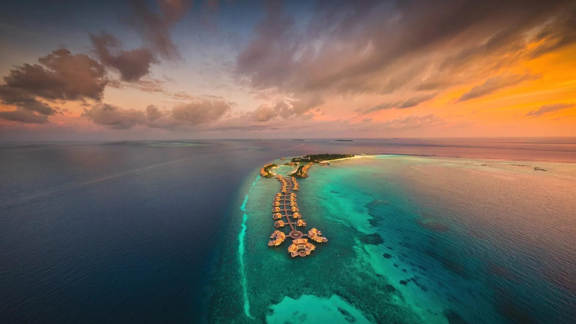 Aerial view at dusk of InterContinental Maldives Maamunagau's overwater villas extending in a line from the island into the Raa Atoll lagoon