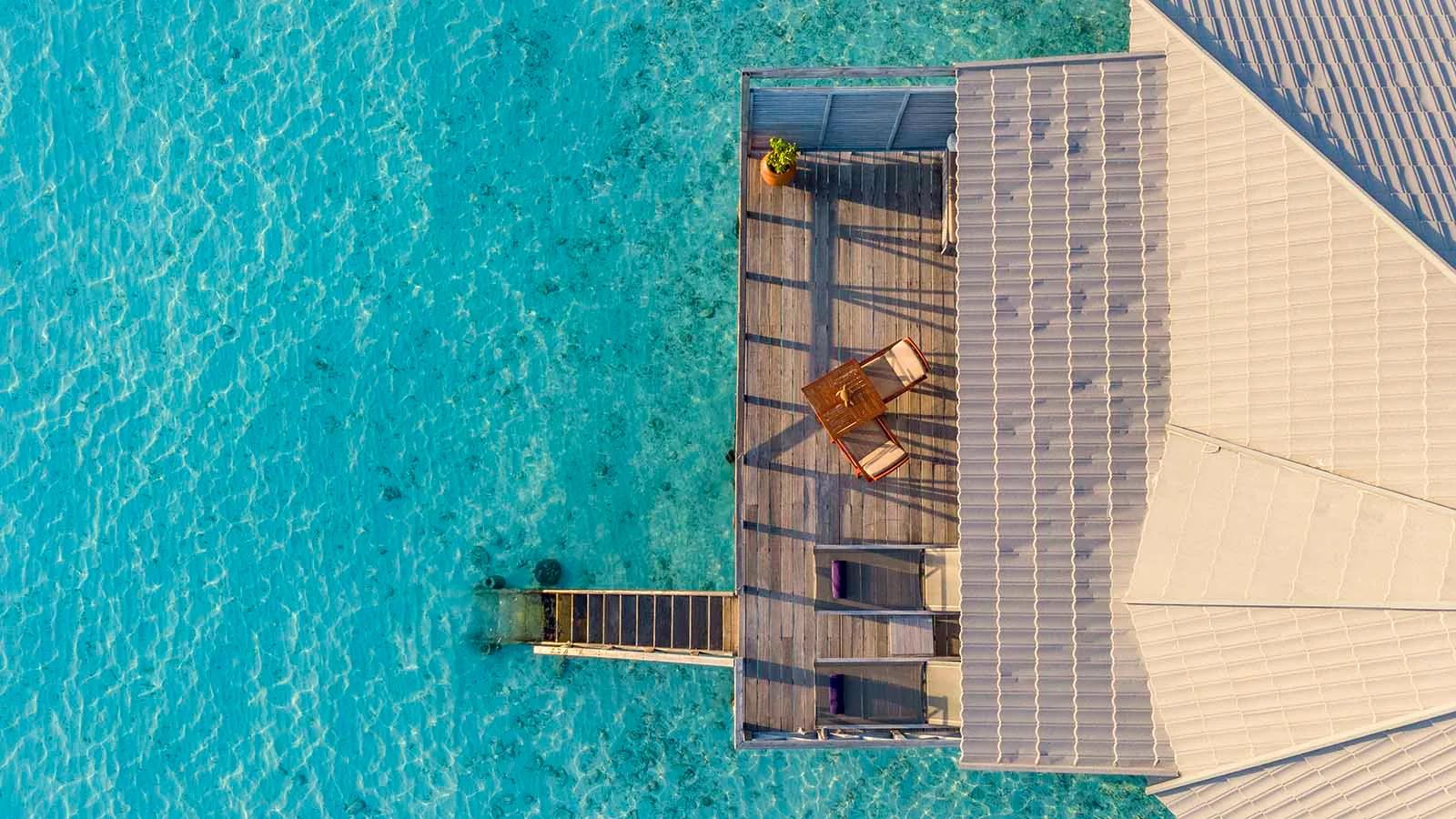A Jacuzzi Water Villa's deck seen directly from above, with a single lounge chair and steps descending into the clear lagoon
