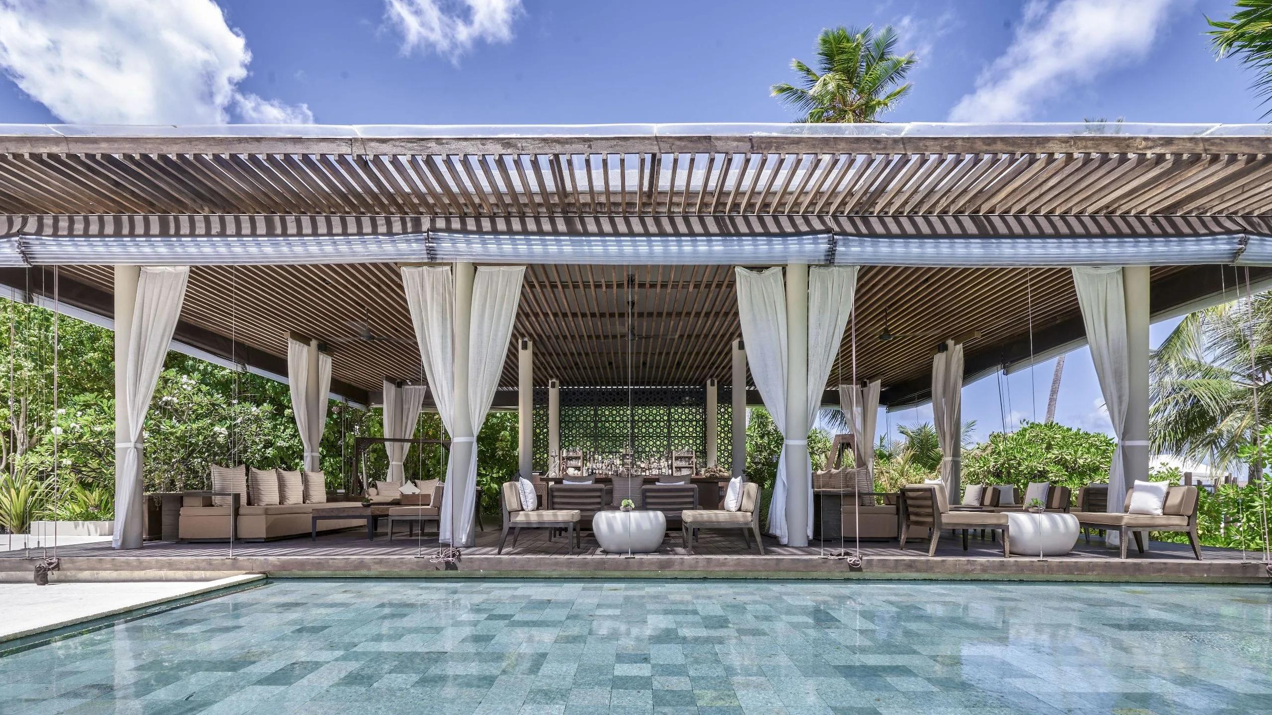 The Bar's shaded lounge seating beside the main pool, white curtains framing the view