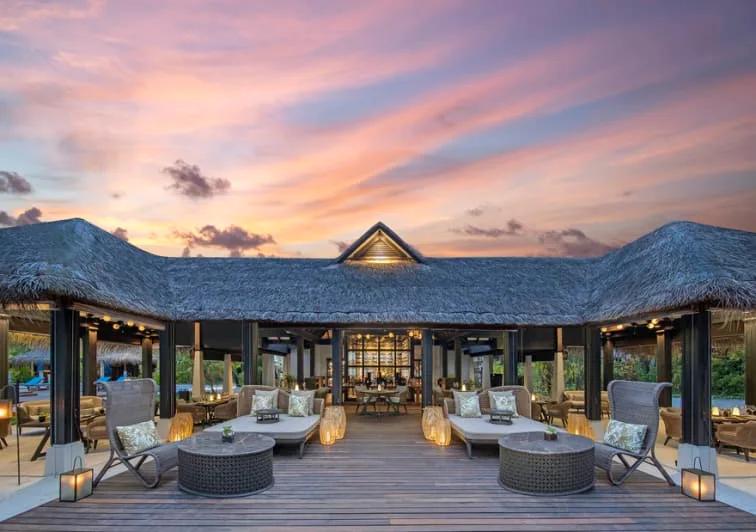 The Living Room's open-air terrace at dusk, thatched roof over lounge seating and lit lanterns as the sky turns pink