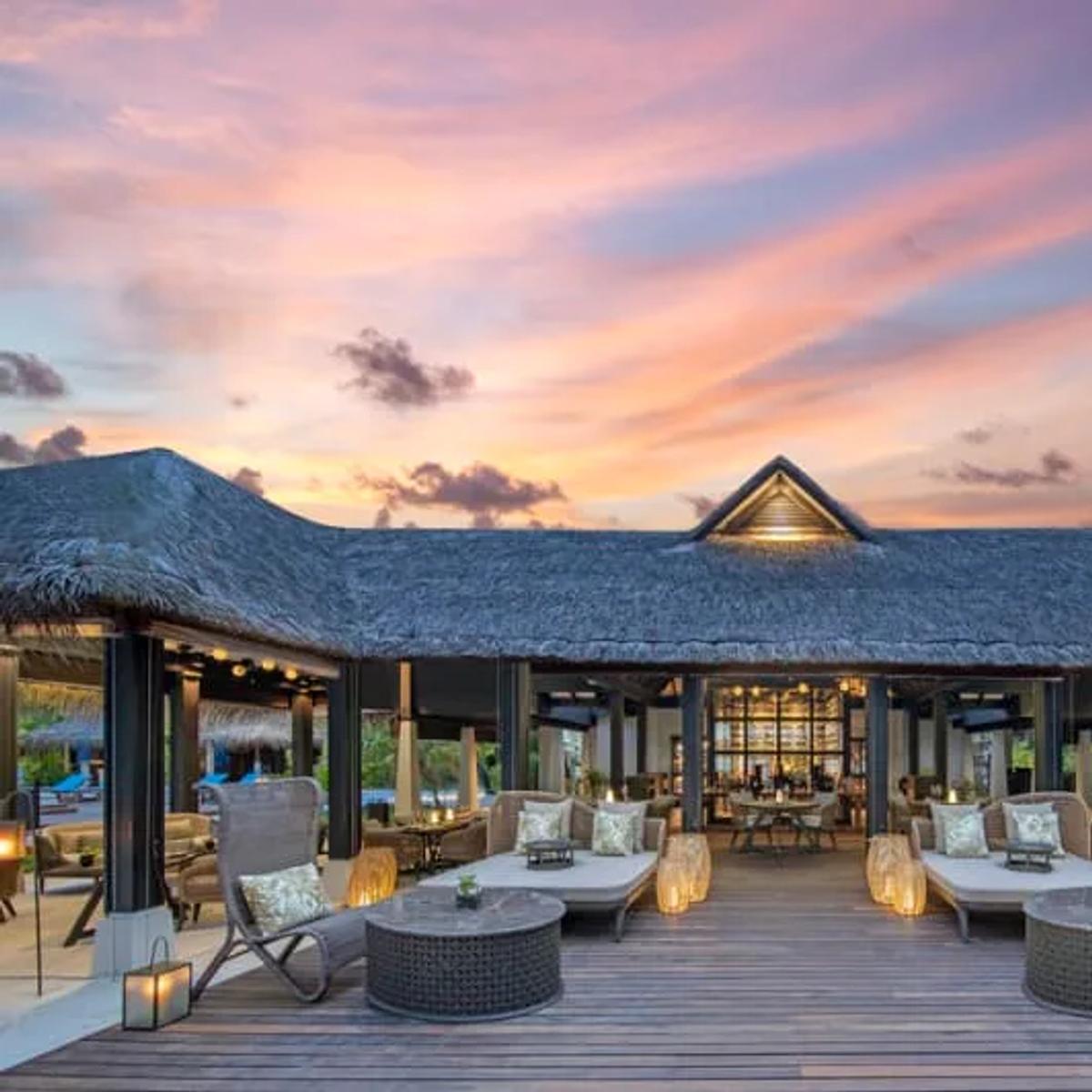 The Living Room's open-air terrace at dusk, thatched roof over lounge seating and lit lanterns as the sky turns pink