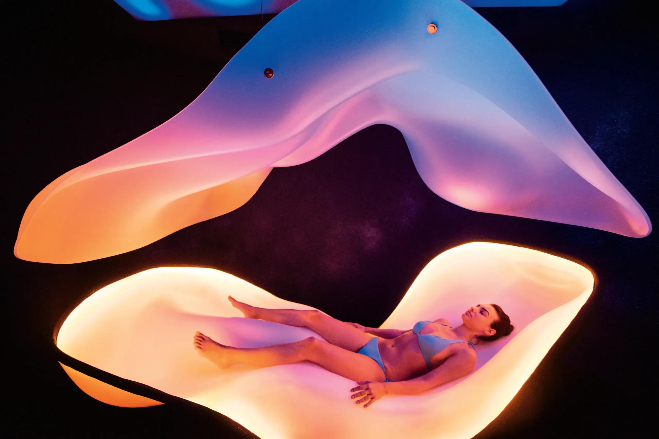 A guest reclines inside the illuminated Dream Pod, a curved overhead canopy glowing pink and blue above her