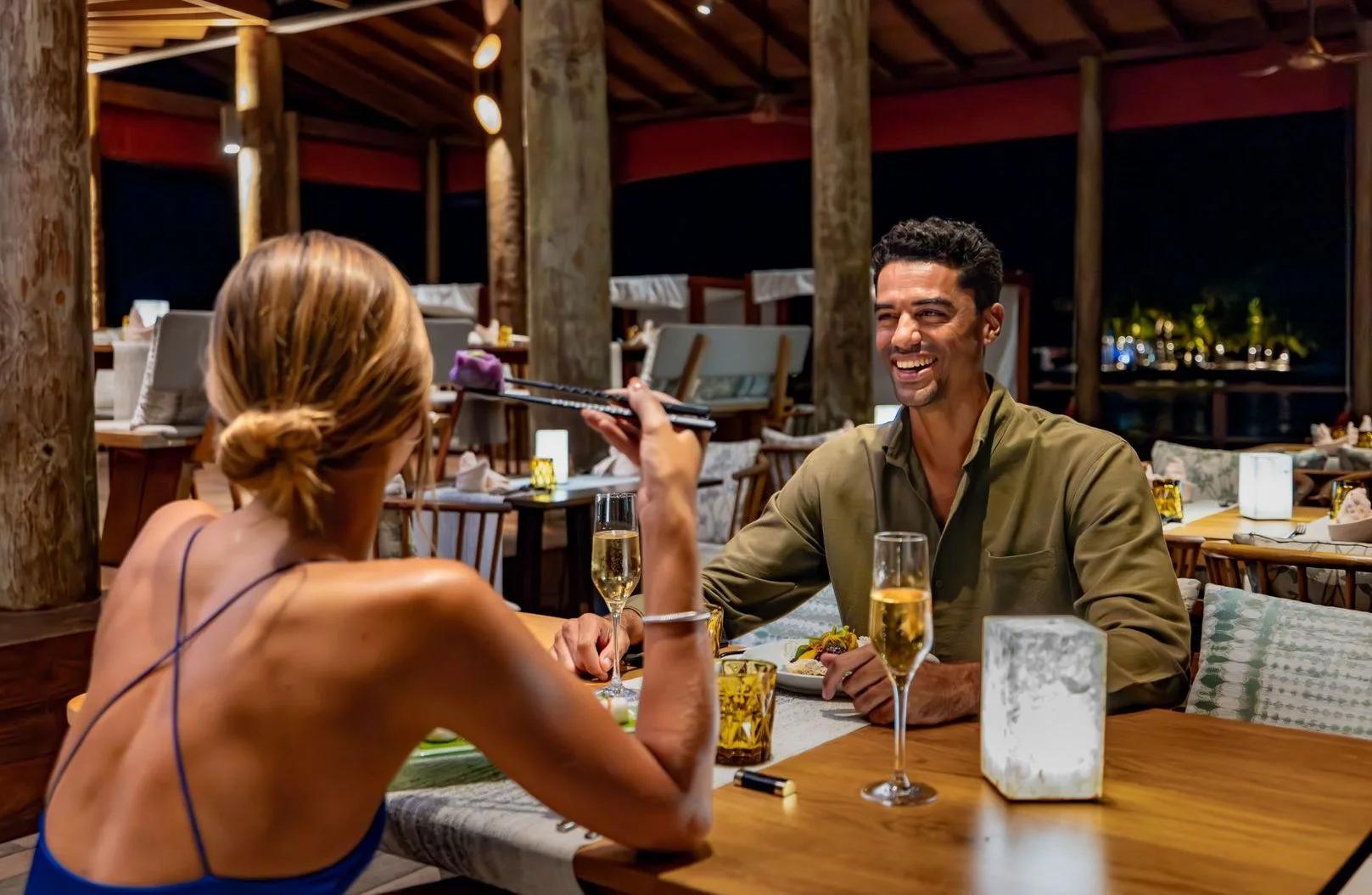 A couple eating with chopsticks at a night-time table beneath a wooden pavilion roof, the lit shoreline visible beyond