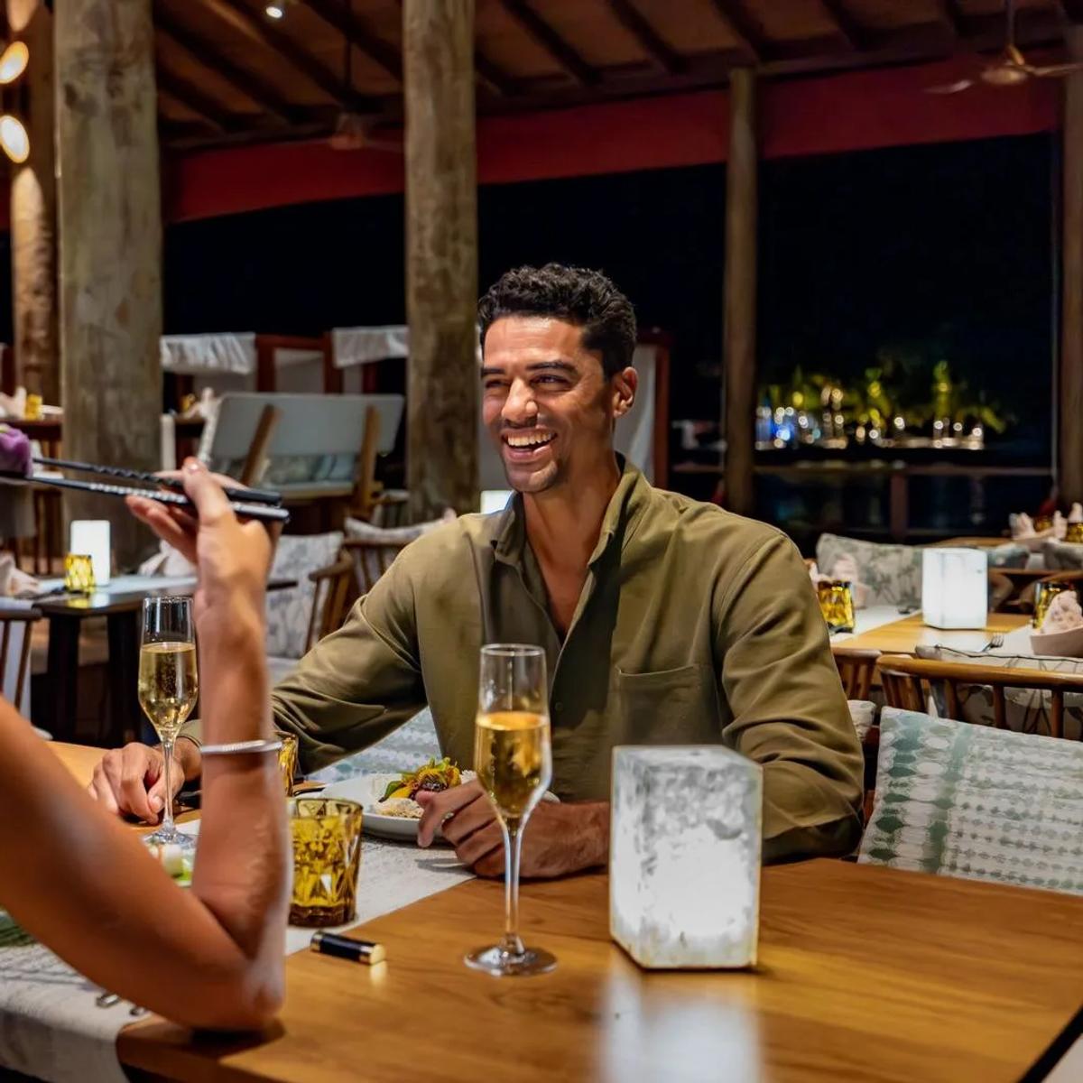 A couple eating with chopsticks at a night-time table beneath a wooden pavilion roof, the lit shoreline visible beyond