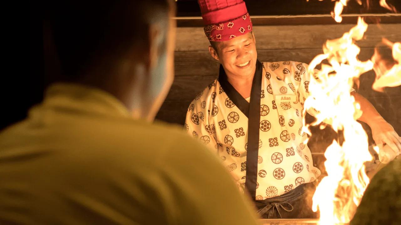 A chef in a happi coat smiling beside open flames at Umami's grill station