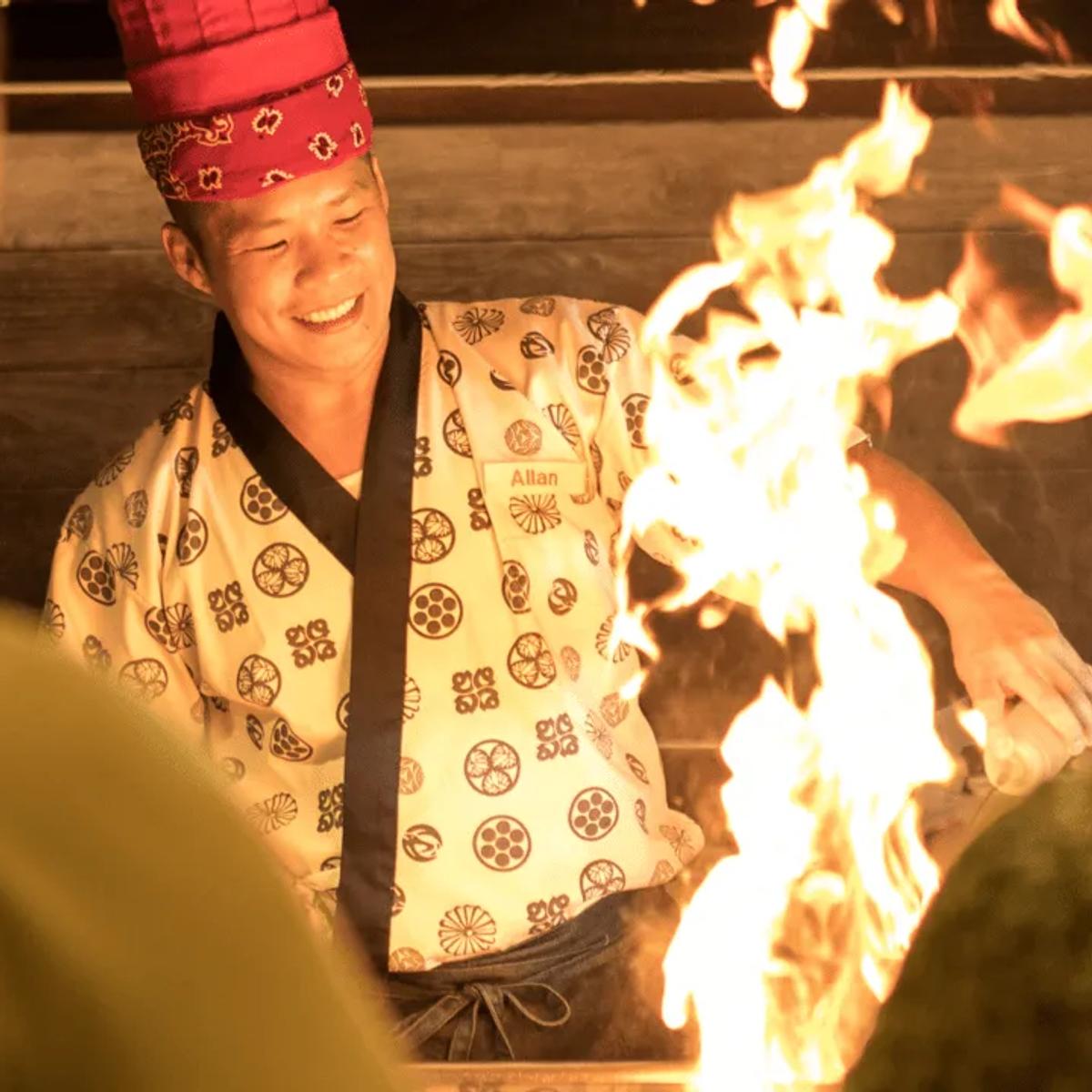 A chef in a happi coat smiling beside open flames at Umami's grill station