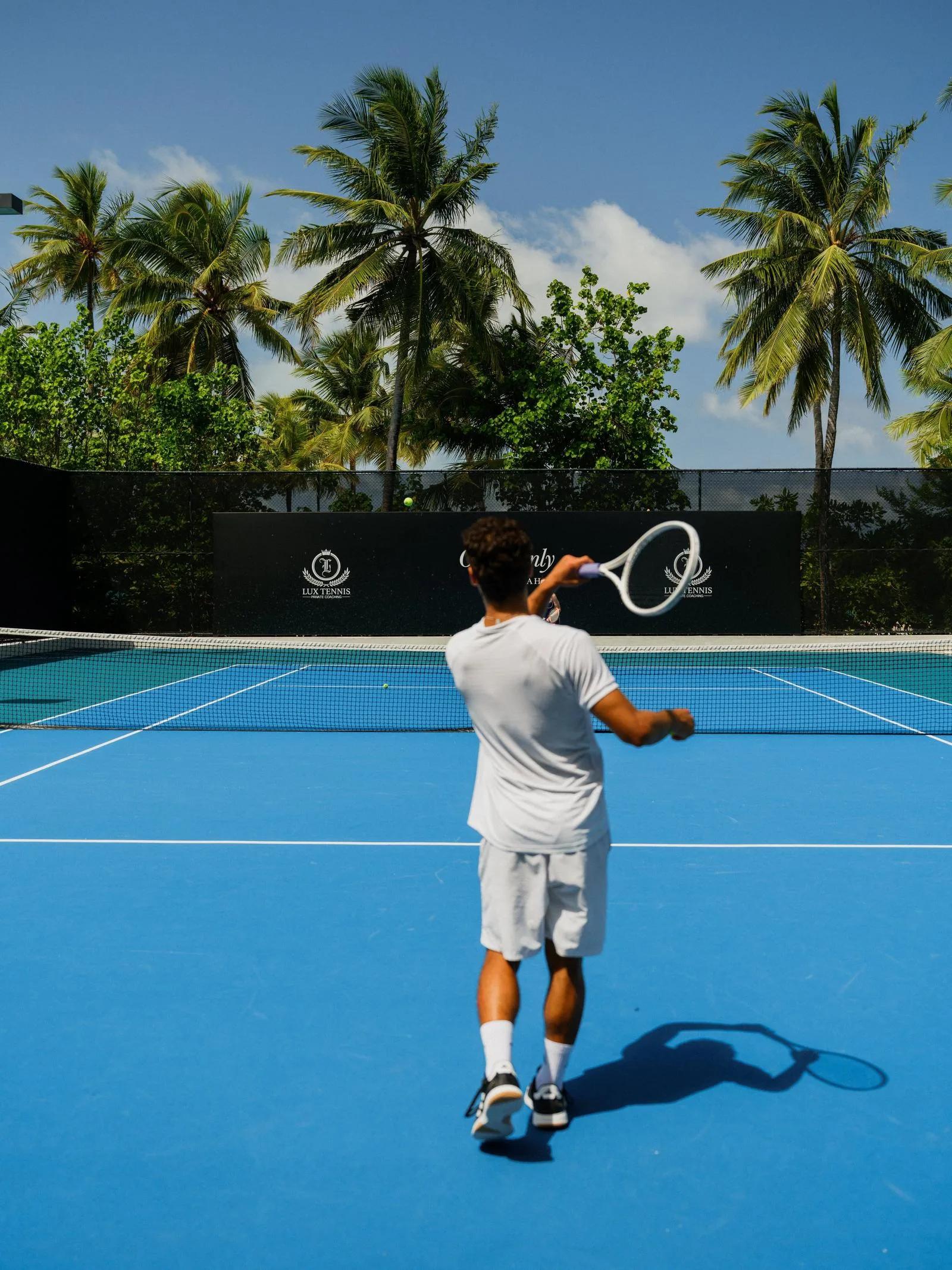 A player mid-swing on one of One&Only Reethi Rah's tennis courts, palm trees behind