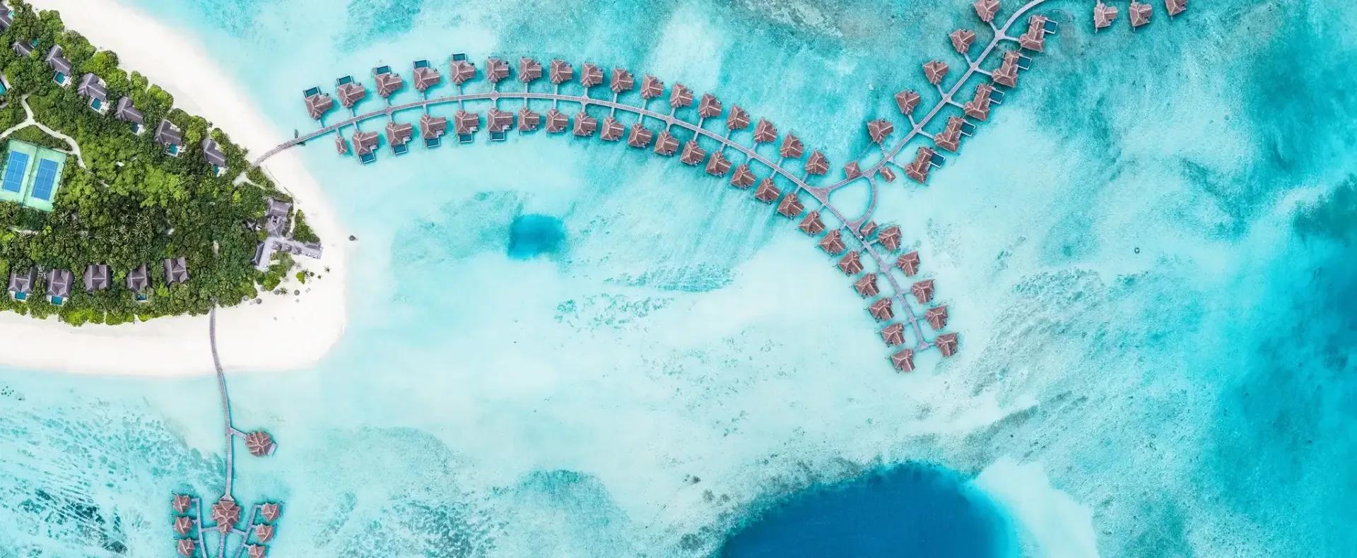 Aerial view of Vakkaru's tree-covered island, a jetty of thatched-roof overwater villas curving out across the reef flat, with a deep blue hole visible in the turquoise lagoon