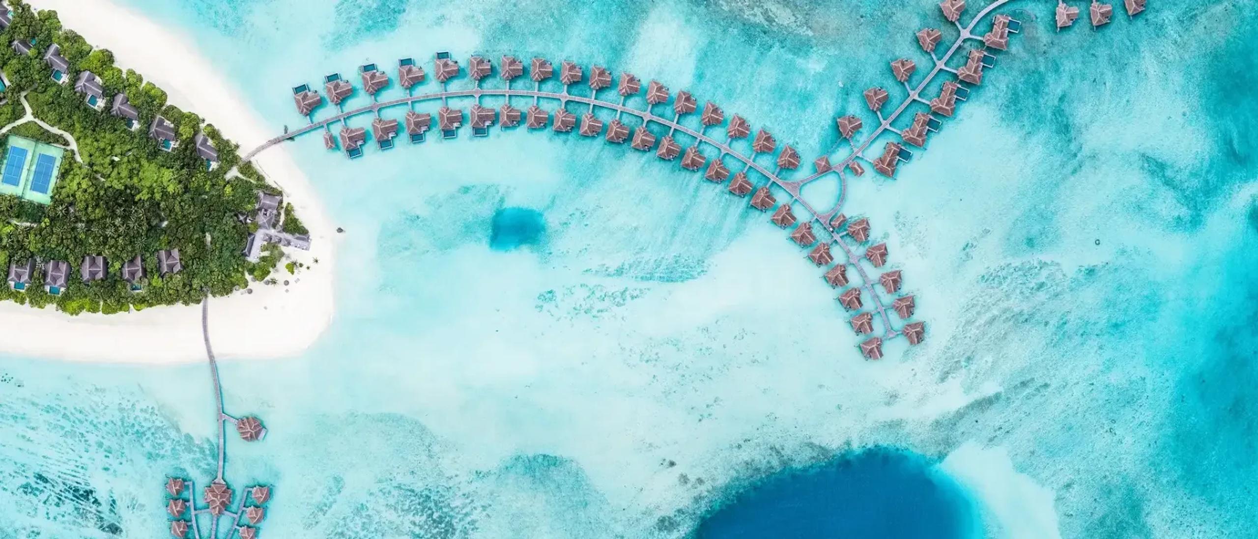 Aerial view of Vakkaru's tree-covered island, a jetty of thatched-roof overwater villas curving out across the reef flat, with a deep blue hole visible in the turquoise lagoon