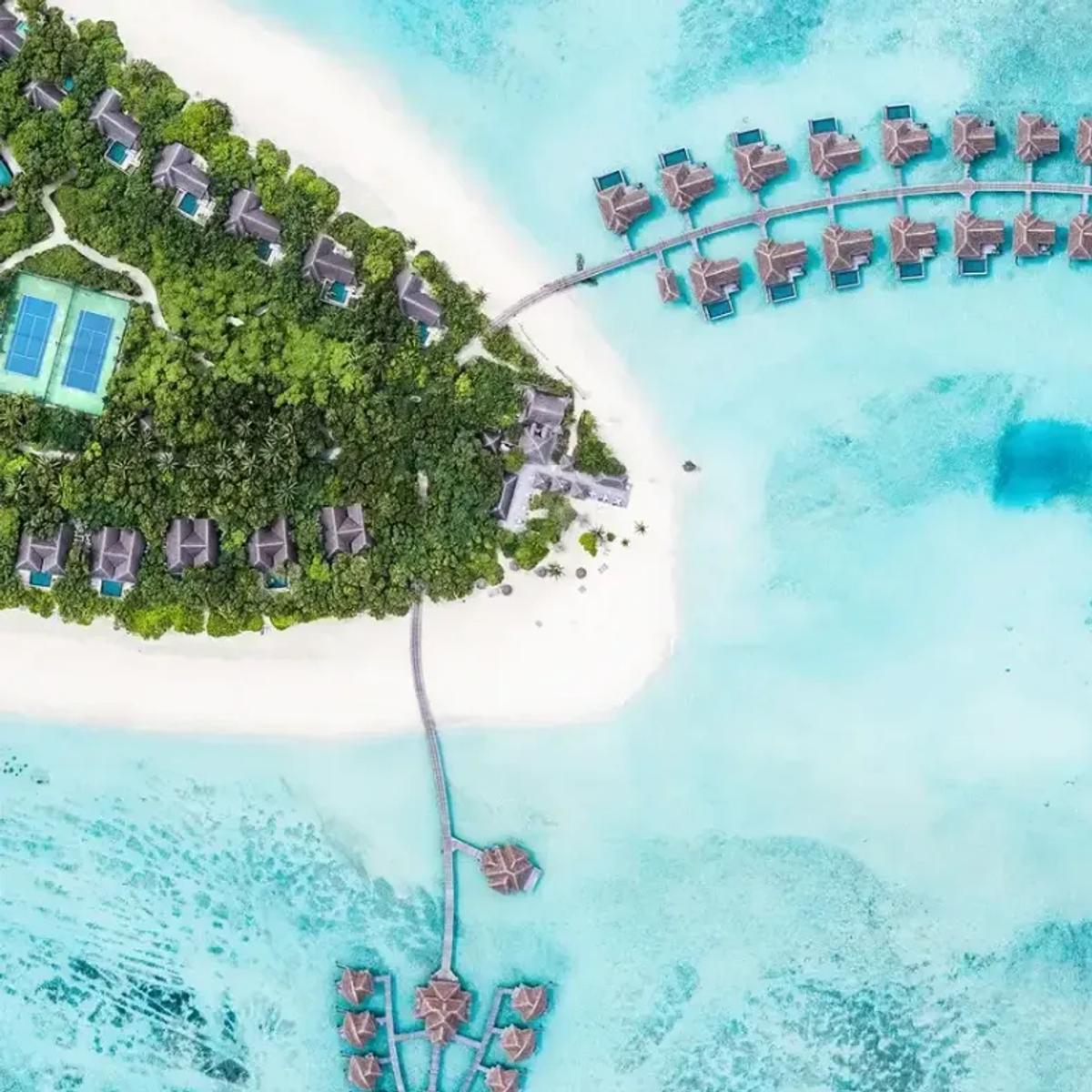 Aerial view of Vakkaru's tree-covered island, a jetty of thatched-roof overwater villas curving out across the reef flat, with a deep blue hole visible in the turquoise lagoon