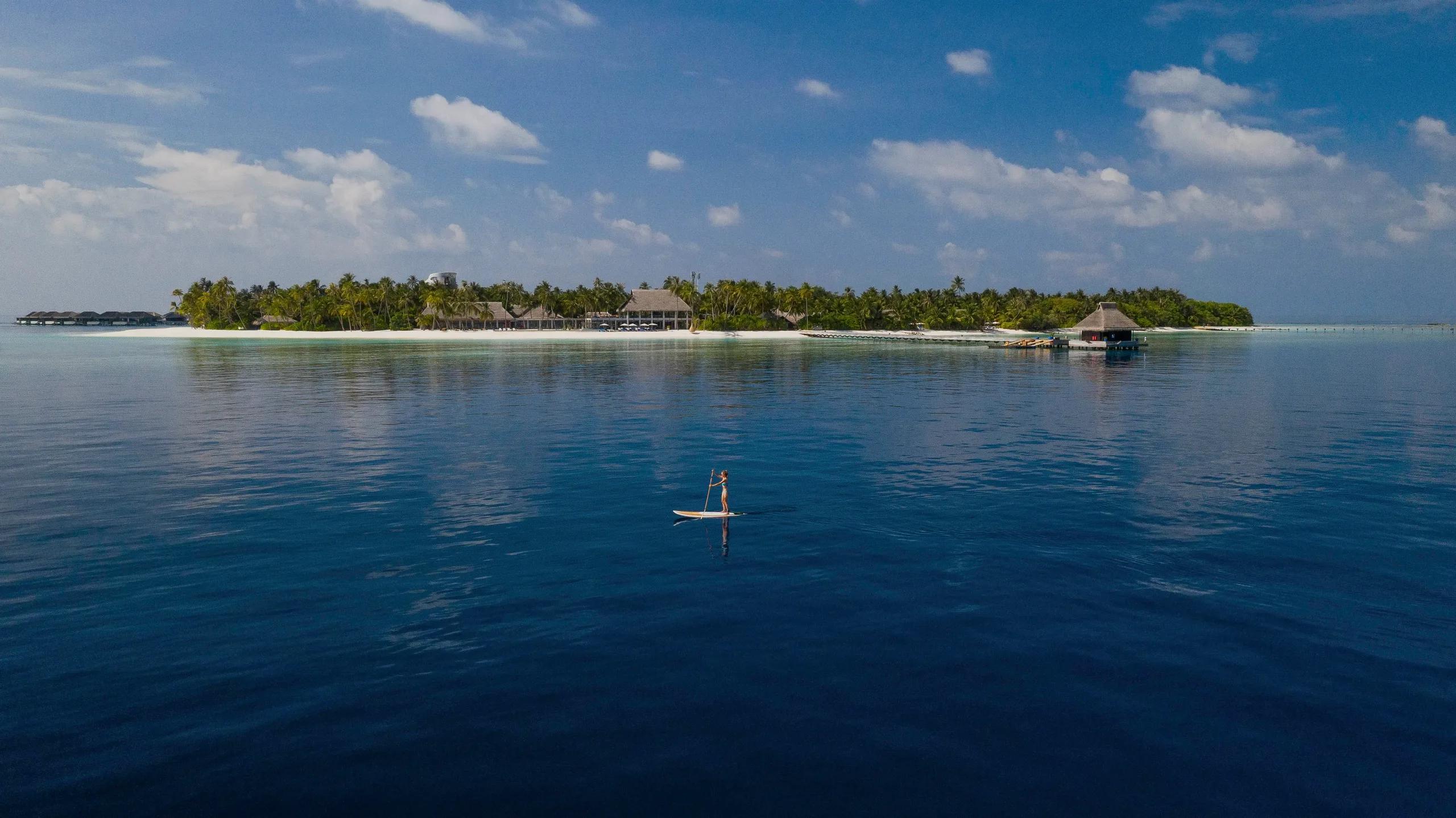 Velaa Private Island seen from the lagoon, overwater villas at left and the main island's thatched roofline behind a paddleboarder crossing calm water