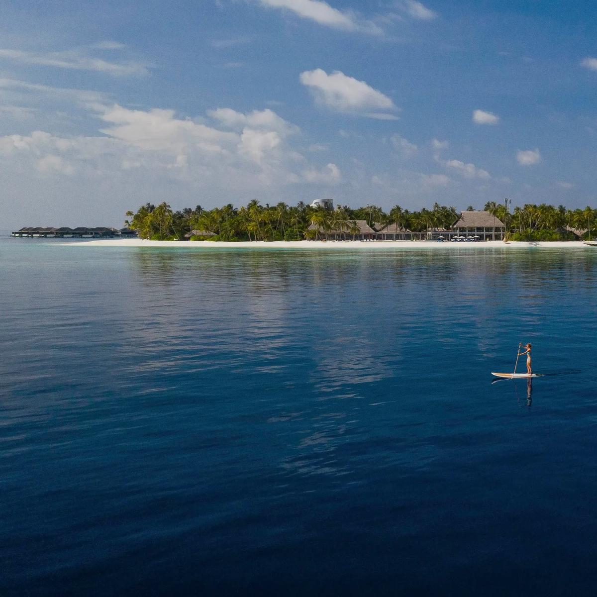 Velaa Private Island seen from the lagoon, overwater villas at left and the main island's thatched roofline behind a paddleboarder crossing calm water