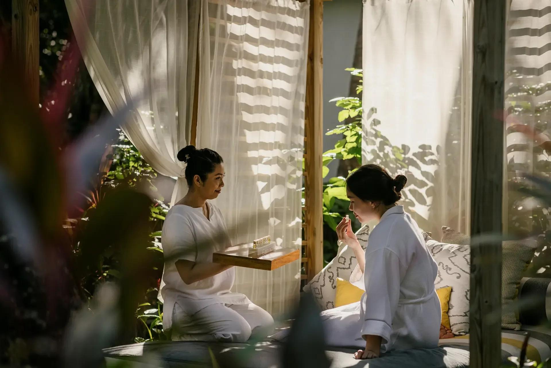 A LIME Wellbeing therapist presenting a tray of oils to a robed guest in a garden lounge