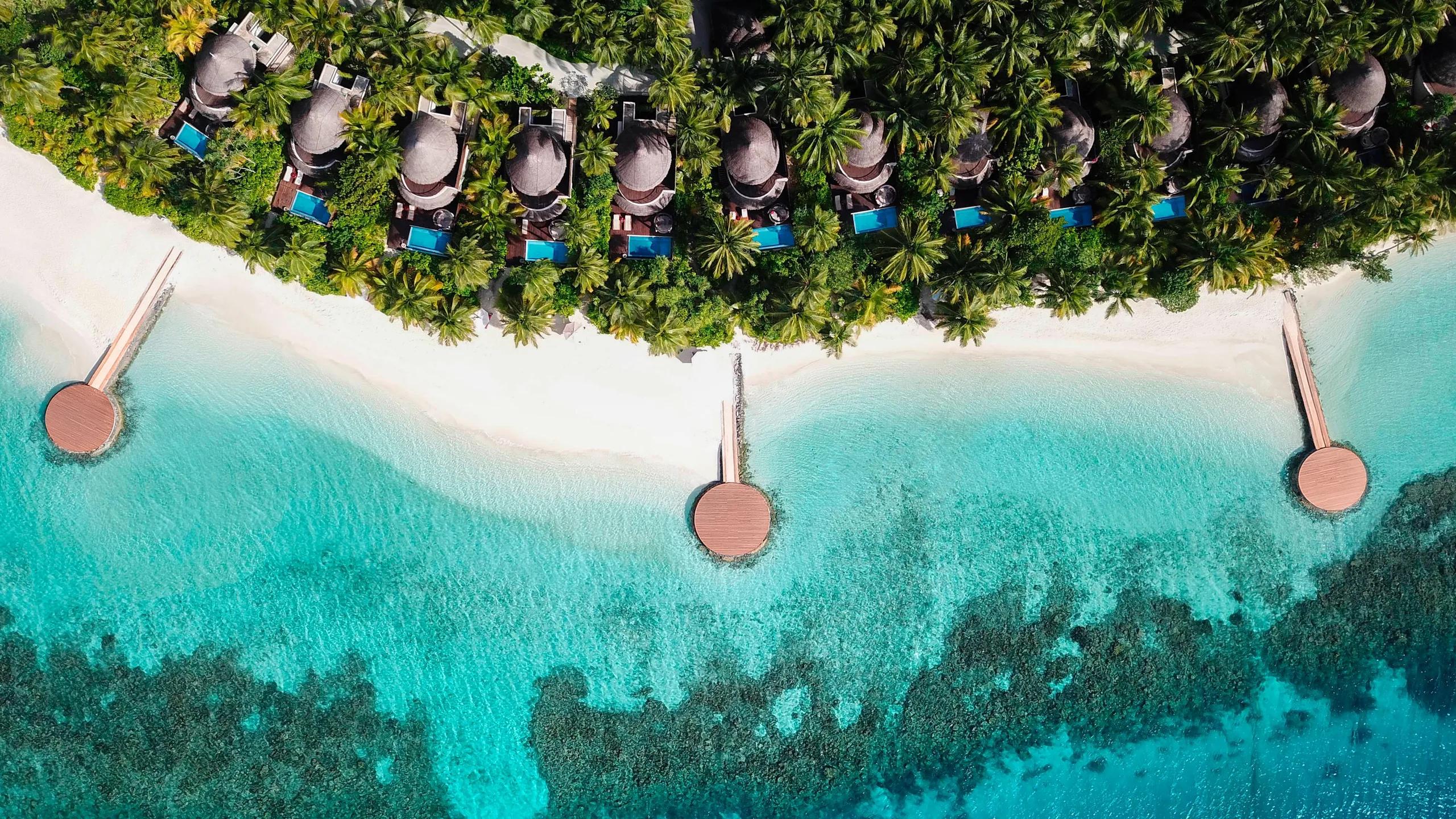 Aerial view of a row of beachfront villas at W Maldives, each with a private pool among the palms, with round sundeck platforms reaching out over the reef on their own jetties