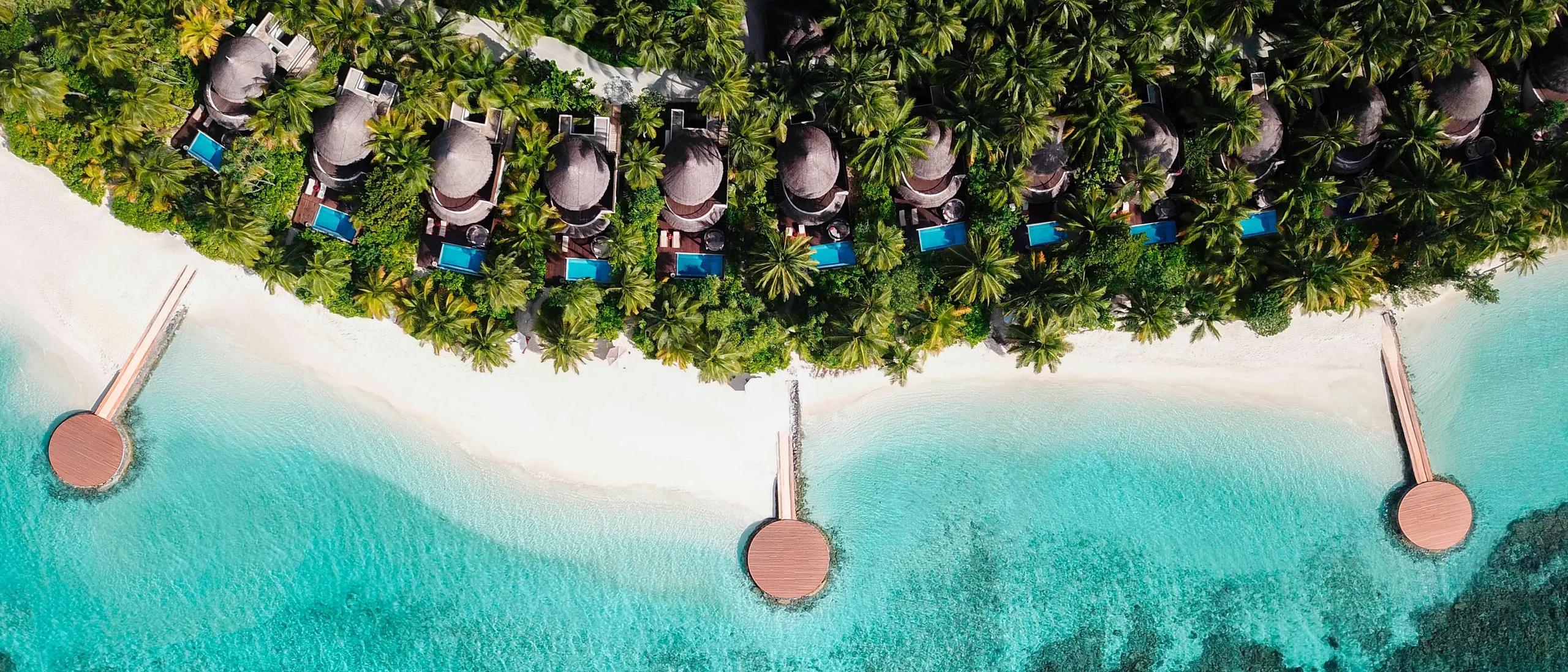 Aerial view of a row of beachfront villas at W Maldives, each with a private pool among the palms, with round sundeck platforms reaching out over the reef on their own jetties