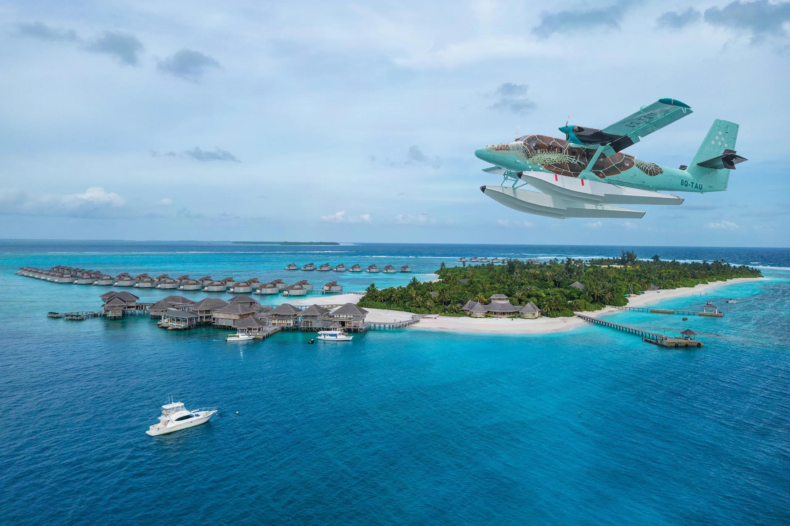 A seaplane on final approach over Olhuveli Island, overwater villas and a moored yacht visible in the lagoon below